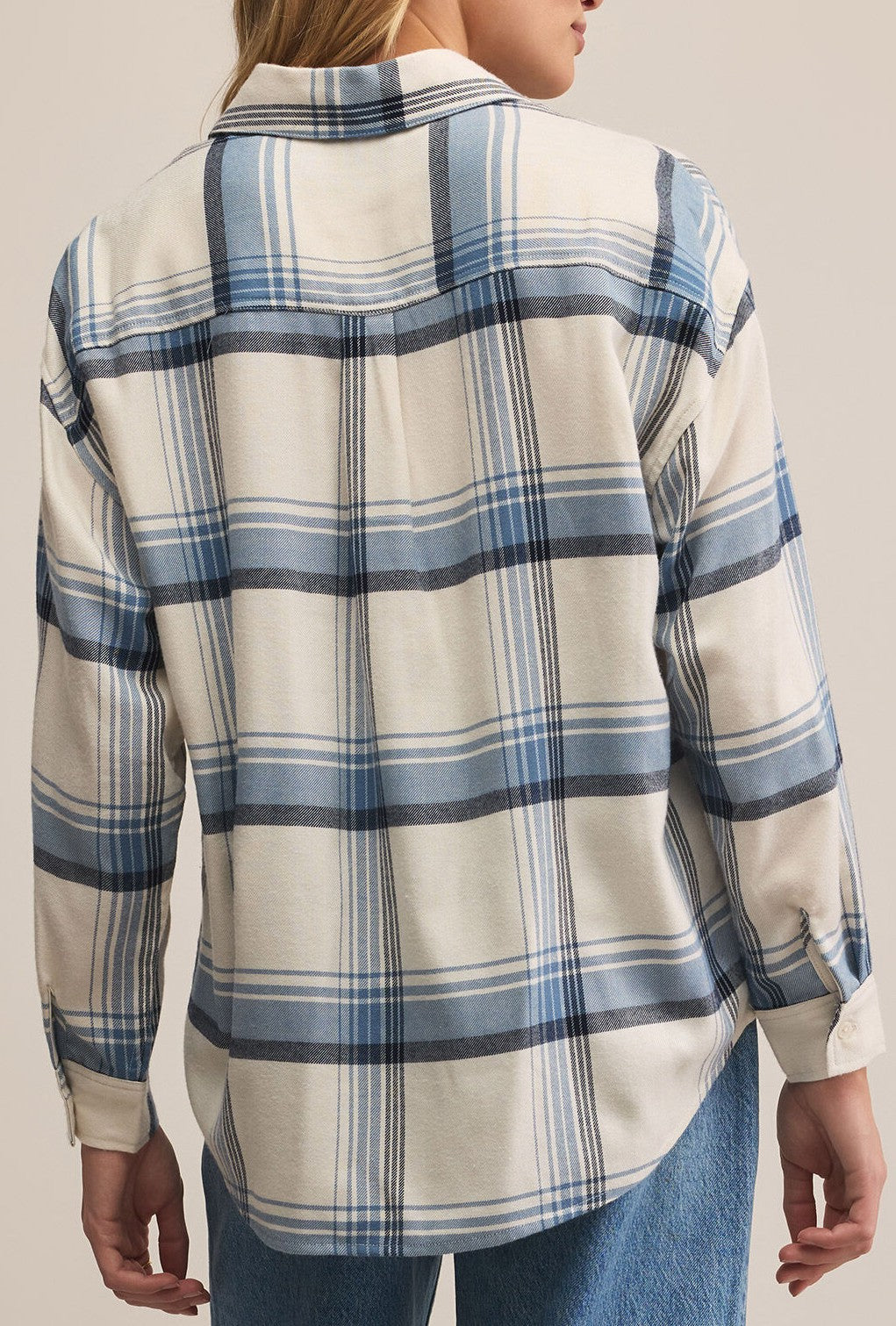 A person is shown from the back wearing the Z Supply River Plaid Button Up In Vintage Indigo, a cream and blue plaid long-sleeve shirt with a relaxed fit, paired with blue jeans. The background is plain and neutral.