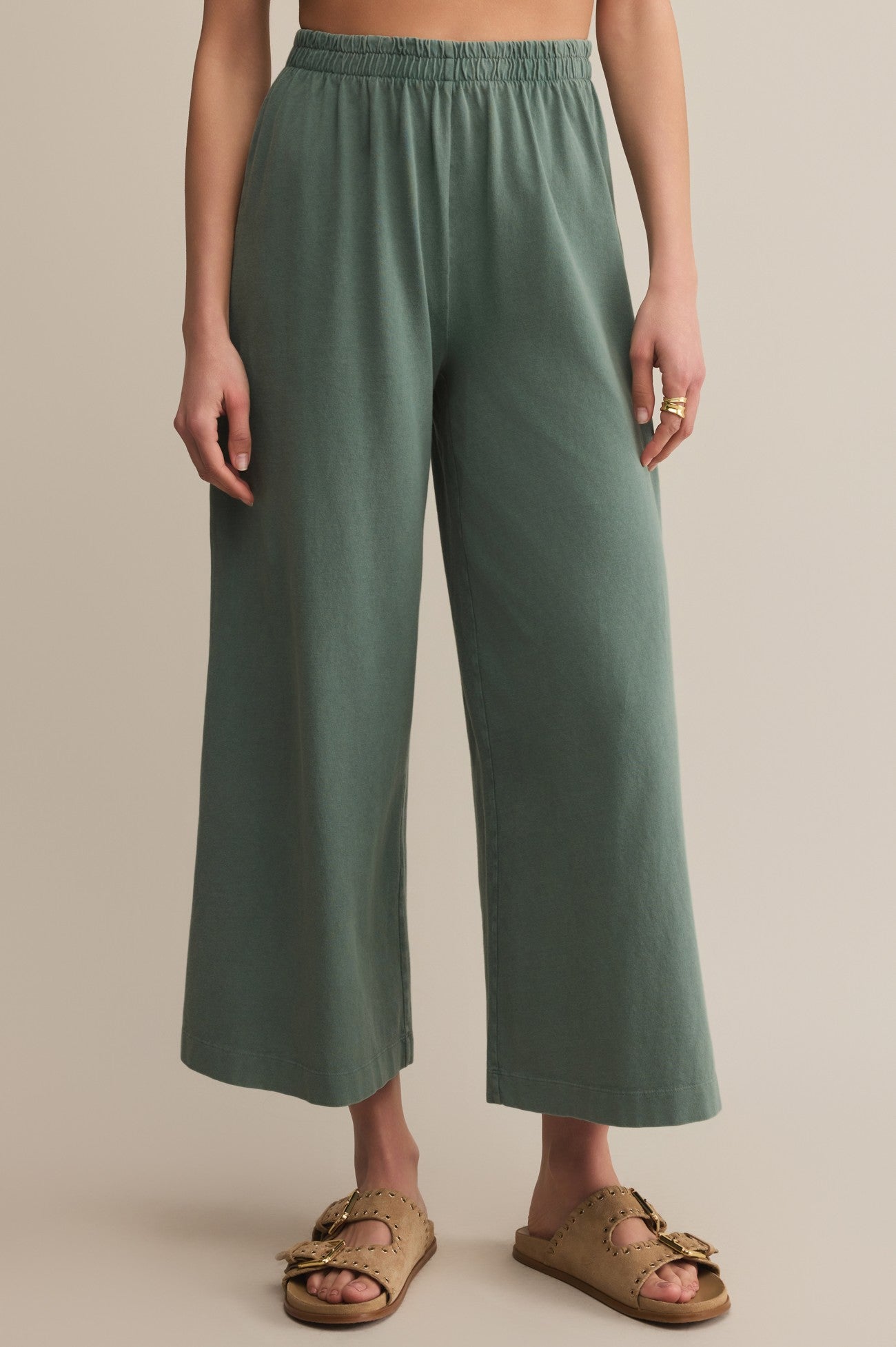 Someone in Z Supply's Scout Jersey Crop Flare Pant in sage green with a relaxed fit and elastic waistband, paired with tan buckled slip-on sandals, stands on a light floor against a beige background.