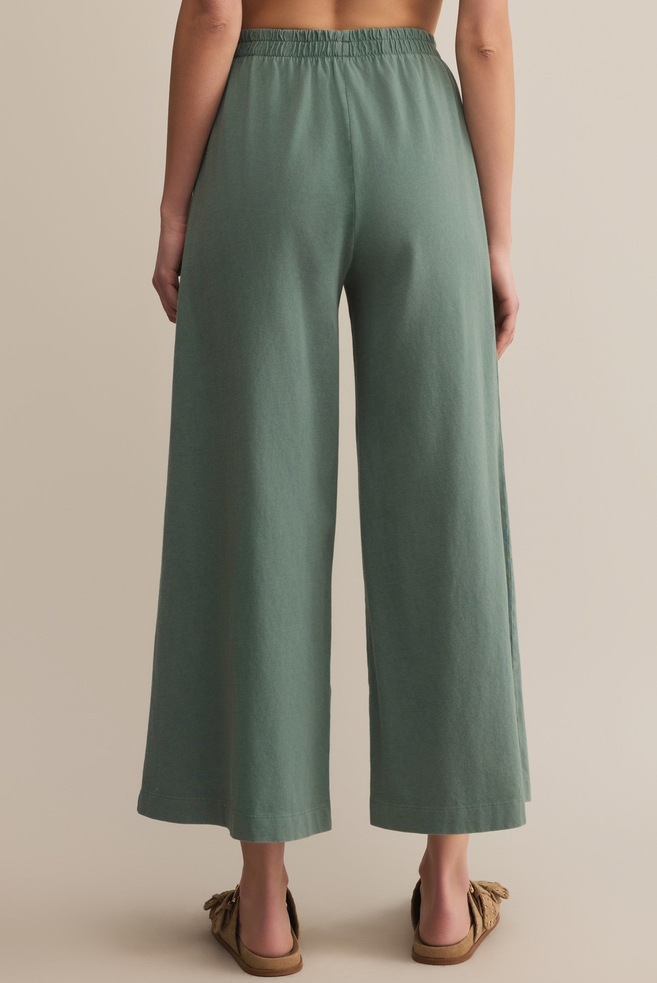 A person is seen from behind wearing Z Supply’s Scout Jersey Crop Flare Pant in sage green—a relaxed fit, cotton jersey with a wide flare and elastic waistband—paired with brown sandals against a beige background.