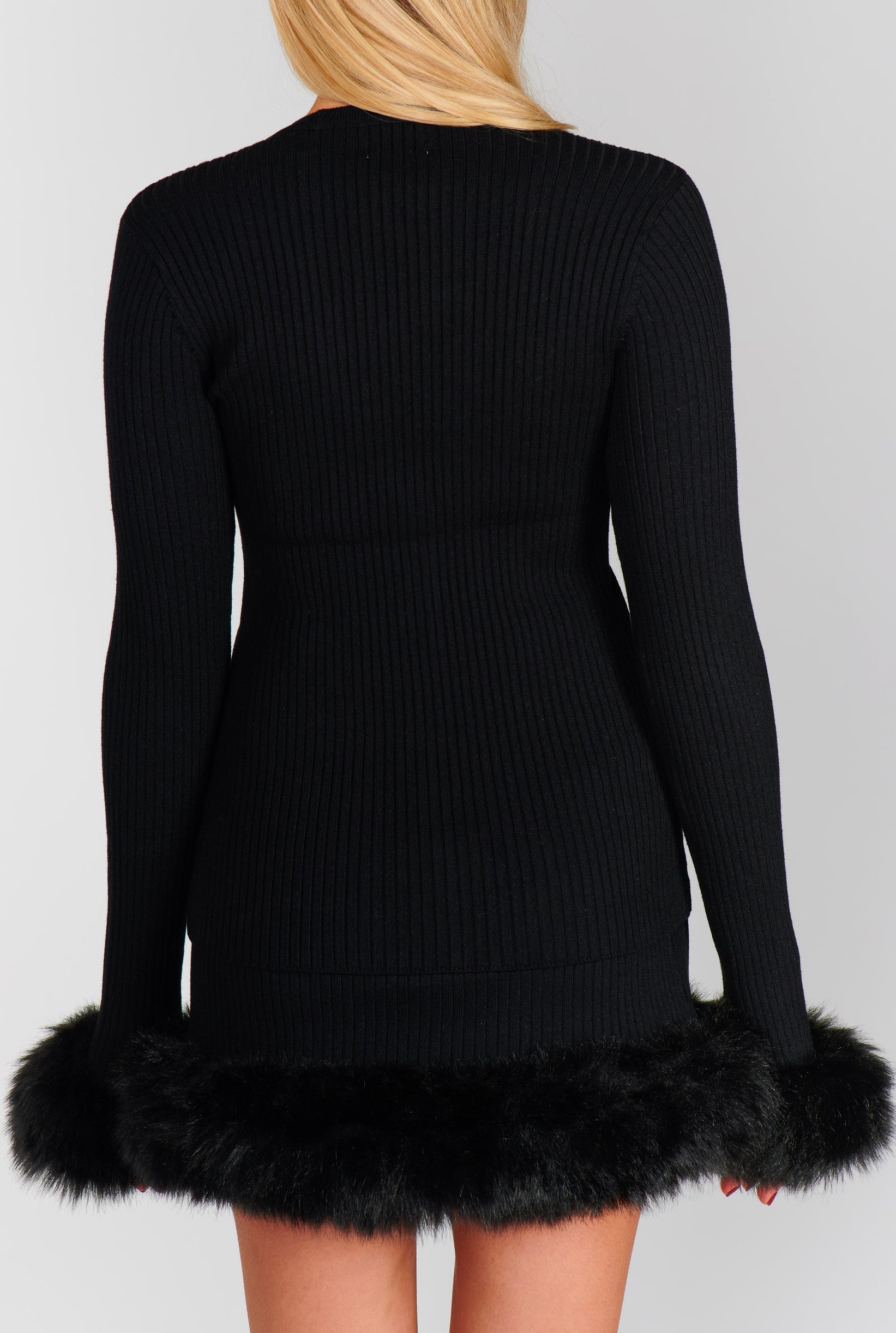 A woman with long blonde hair is shown from the back wearing the Fran Mini Skirt by Show Me Your Mumu—a black, ribbed, long-sleeve dress with black faux fur trim—styled against a plain light gray background.