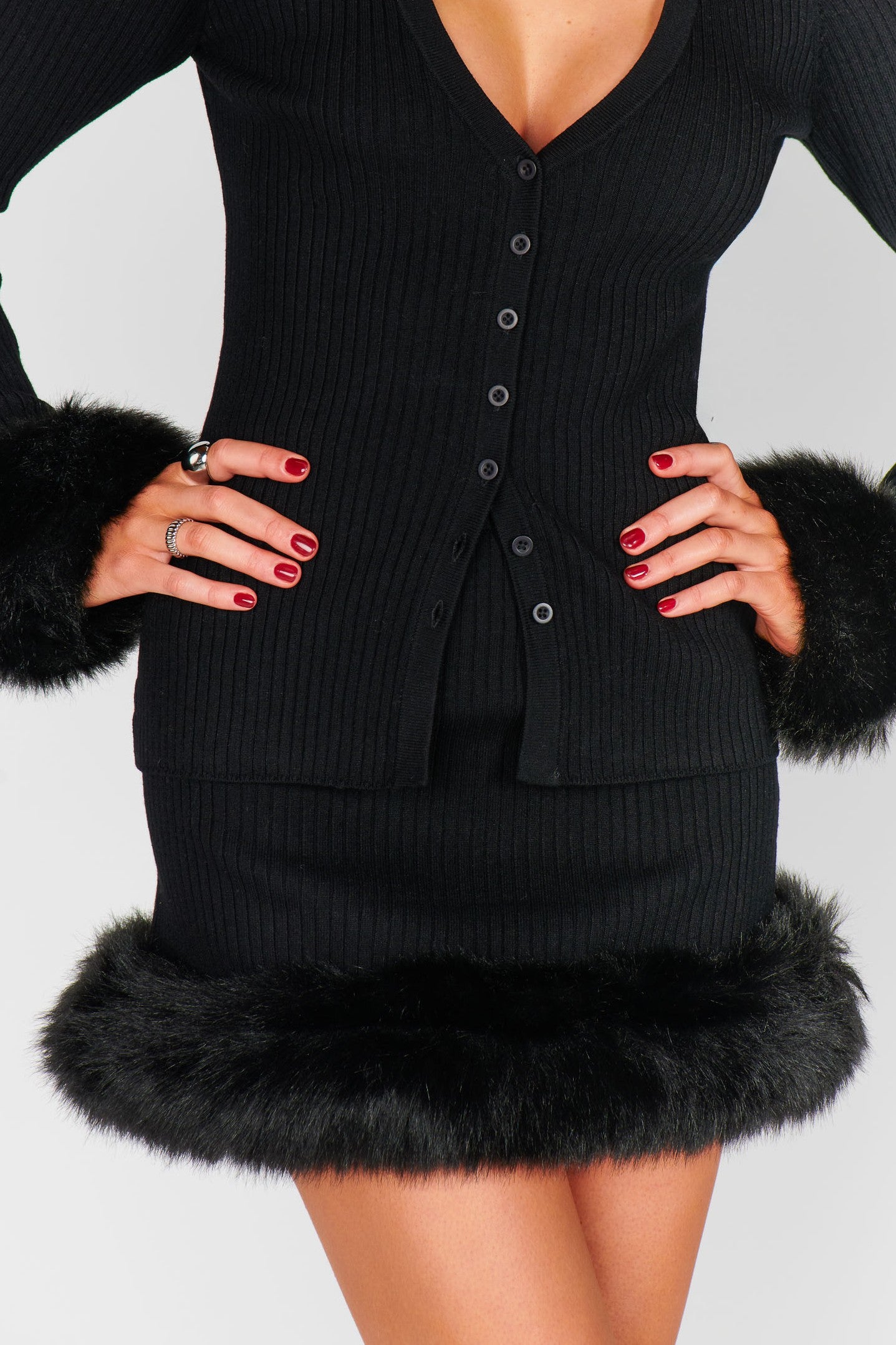 A person wearing the Fran Mini Skirt by Show Me Your Mumu, a fitted faux fur mini skirt with black trim on cuffs and hem, paired with a black ribbed cardigan, hands on hips, showing off red polished nails and rings.