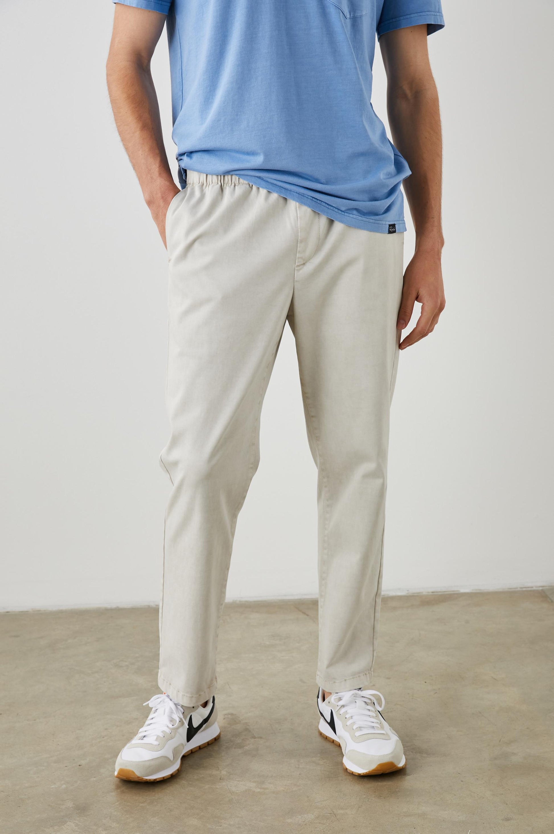 A person wearing light beige Julian Pants by Rails, a blue T-shirt, and white sneakers with black accents stands on a concrete floor against a plain white wall. The person’s face is not visible.