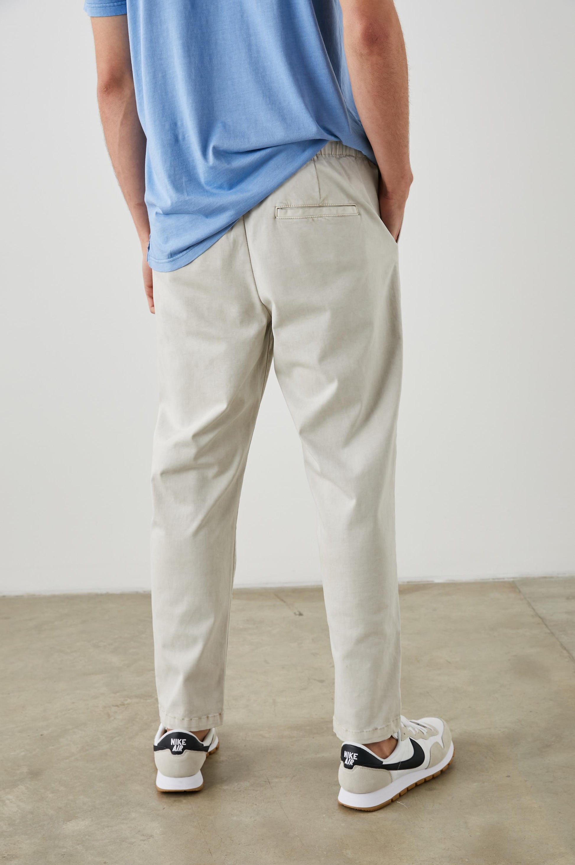 Indoors on a concrete floor, a person stands facing away with hands in pockets. They wear the Rails Julian Pant in light beige, paired with a light blue shirt and white sneakers featuring black accents.