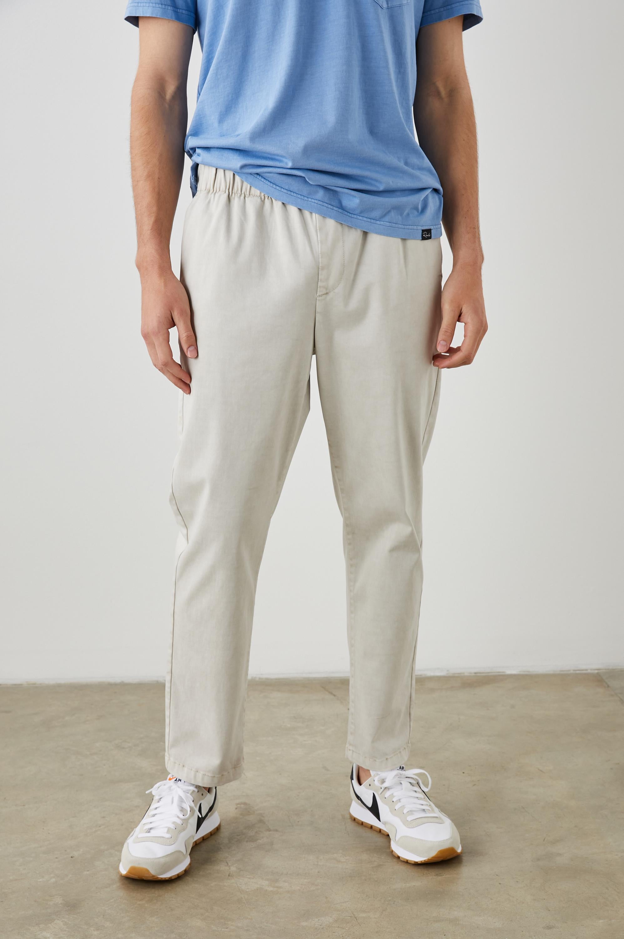 A person stands indoors on a concrete floor, wearing a blue t-shirt, white sneakers with black details, and the Julian Pant by Rails in light beige with an elastic waistband. Their upper face is not visible.