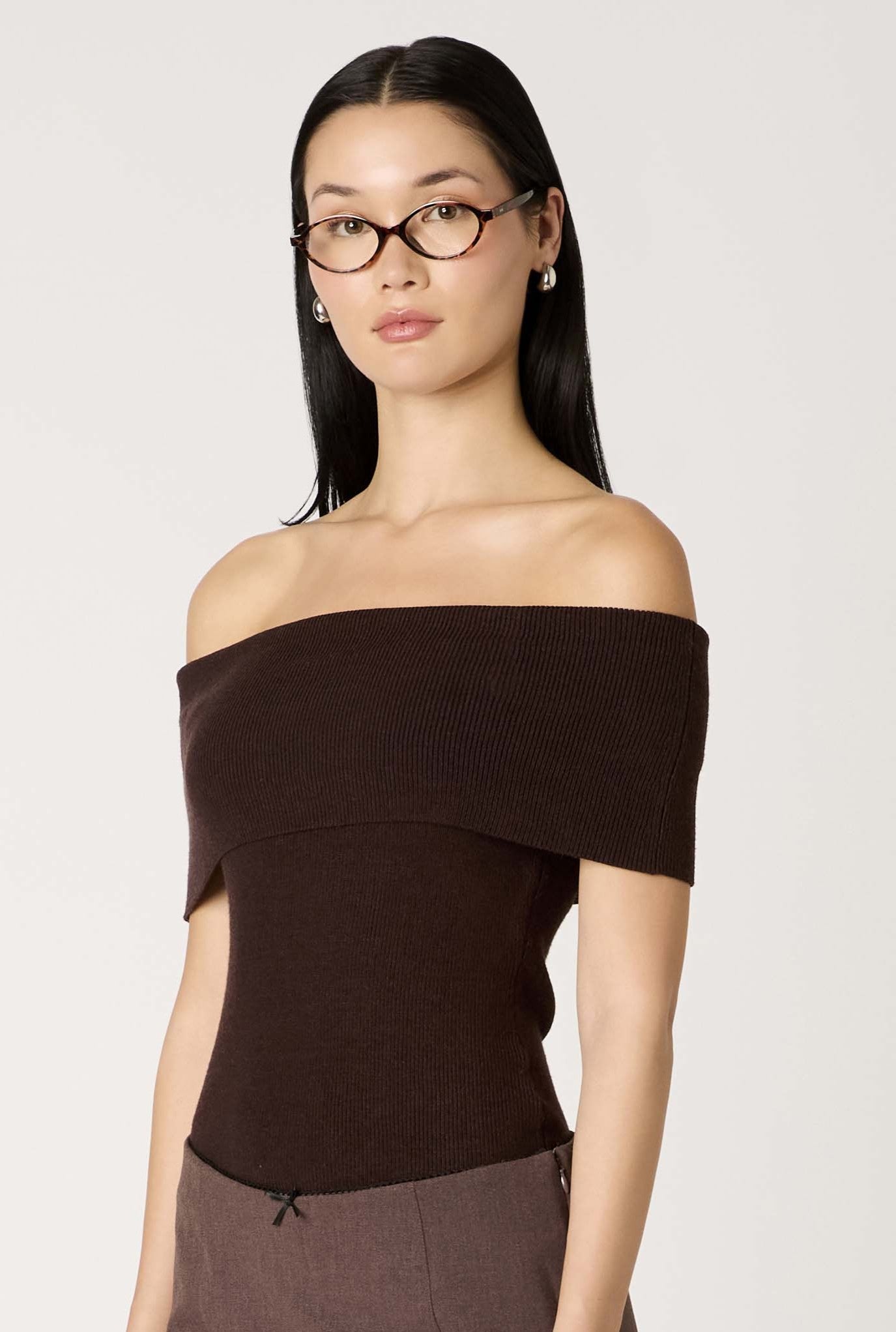 A woman with straight black hair, glasses, stud earrings, and a dark brown Annemarie Sweater by Nia stands against a plain light background, looking forward with a neutral expression.