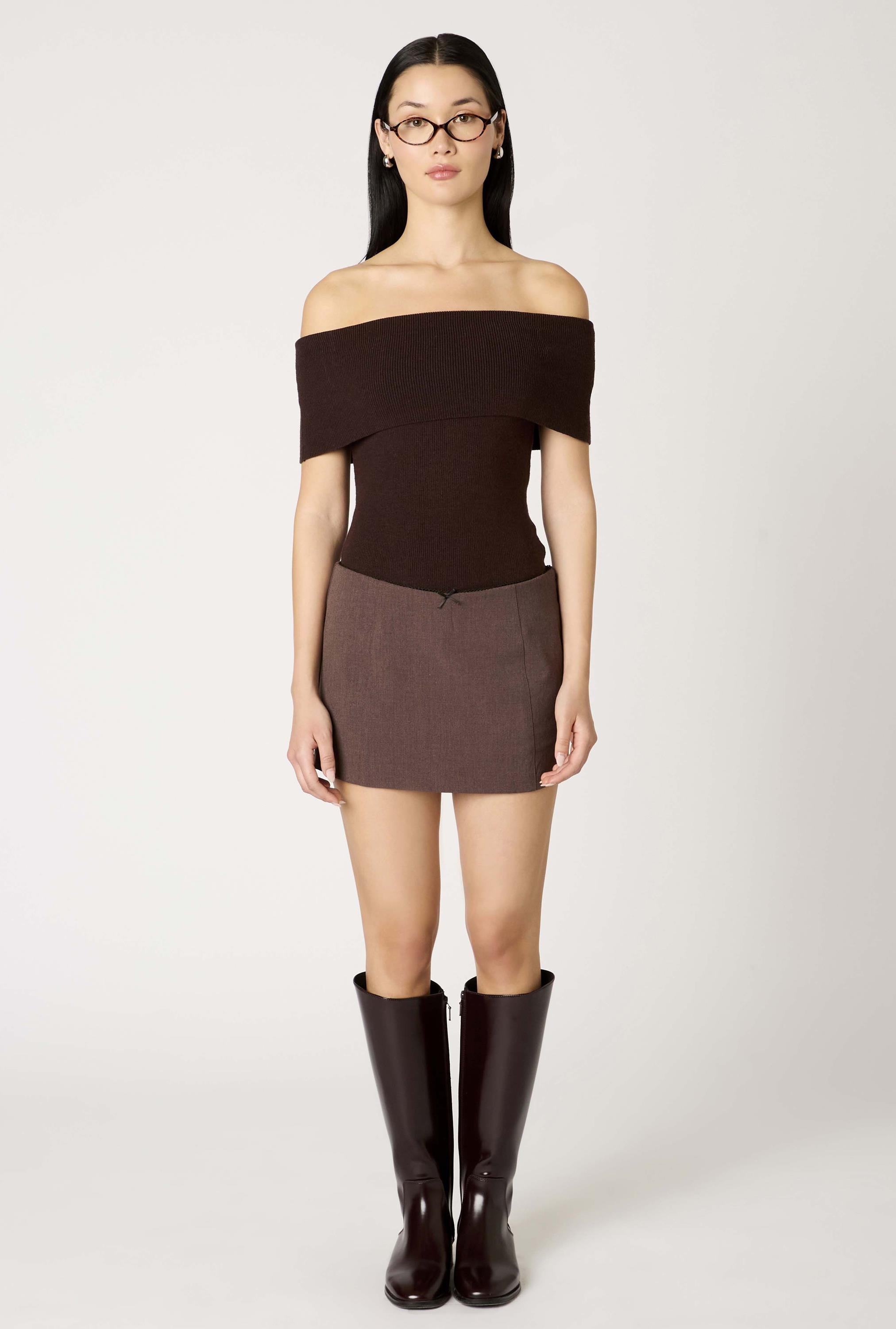 A woman faces forward against a plain light background wearing Nia’s Annemarie Sweater, styled with glasses, a short brown skirt, and knee-high dark boots; her arms are at her sides.
