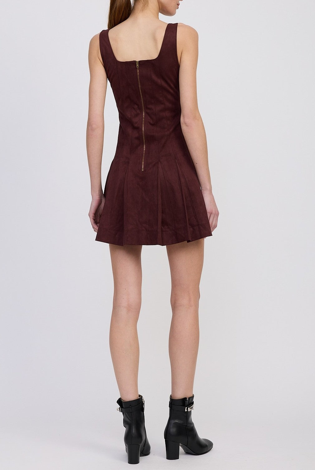 A woman stands facing away, wearing the En Saison Delphine Pleated Mini Dress—a sleeveless, dark maroon mini dress featuring a back zipper and a pleated flared skirt. She pairs it with black buckle ankle boots against a plain, light-colored background.