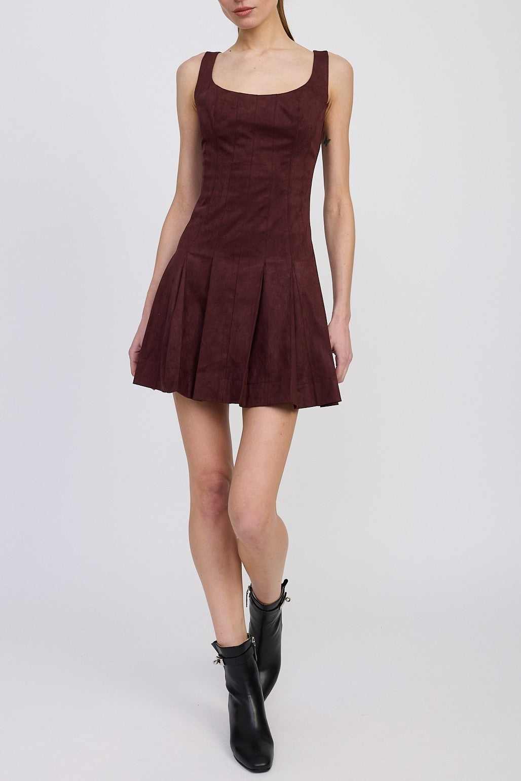 A person wearing the Delphine Pleated Mini Dress by En Saison in burgundy, featuring a square neckline and a pleated flared skirt, paired with black ankle boots, stands against a plain light background.