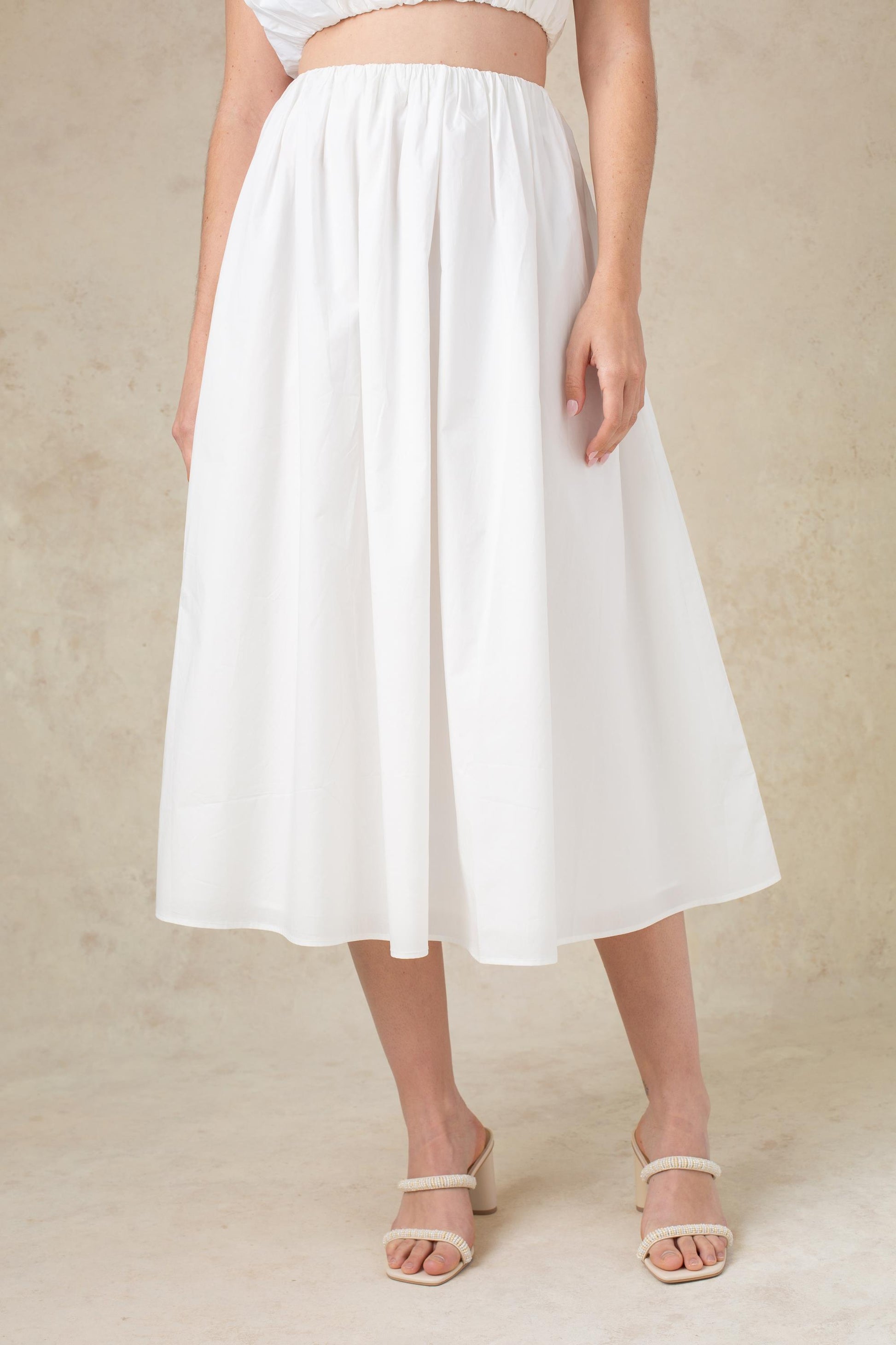 A woman models the Georgie Bari Poplin Skirt—a white, high-waisted midi with a gathered waist—paired with beige heeled sandals against a neutral background. Her face is not shown.
