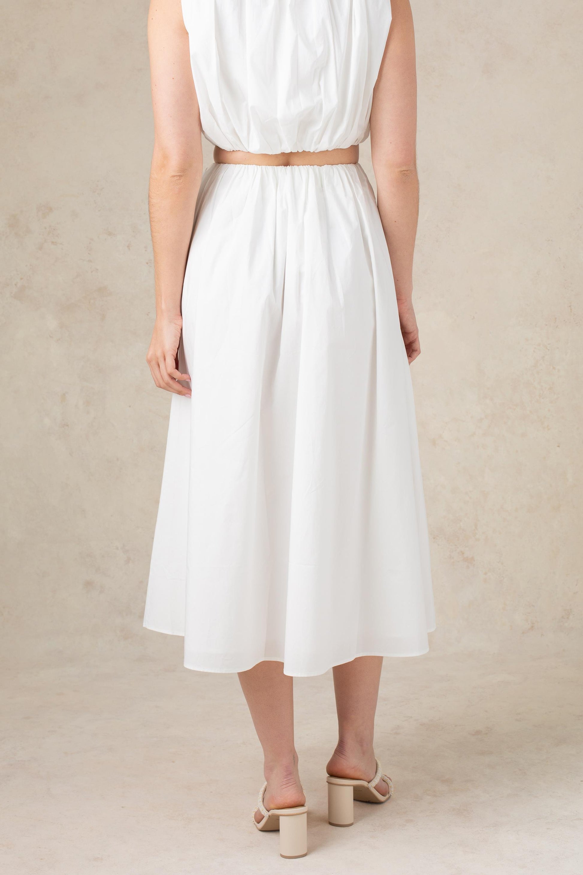A woman stands on a beige surface, facing away from the camera. She is wearing the Georgie Bari Poplin Skirt, a white, calf-length style paired with beige heeled sandals. Her arms hang naturally by her sides.