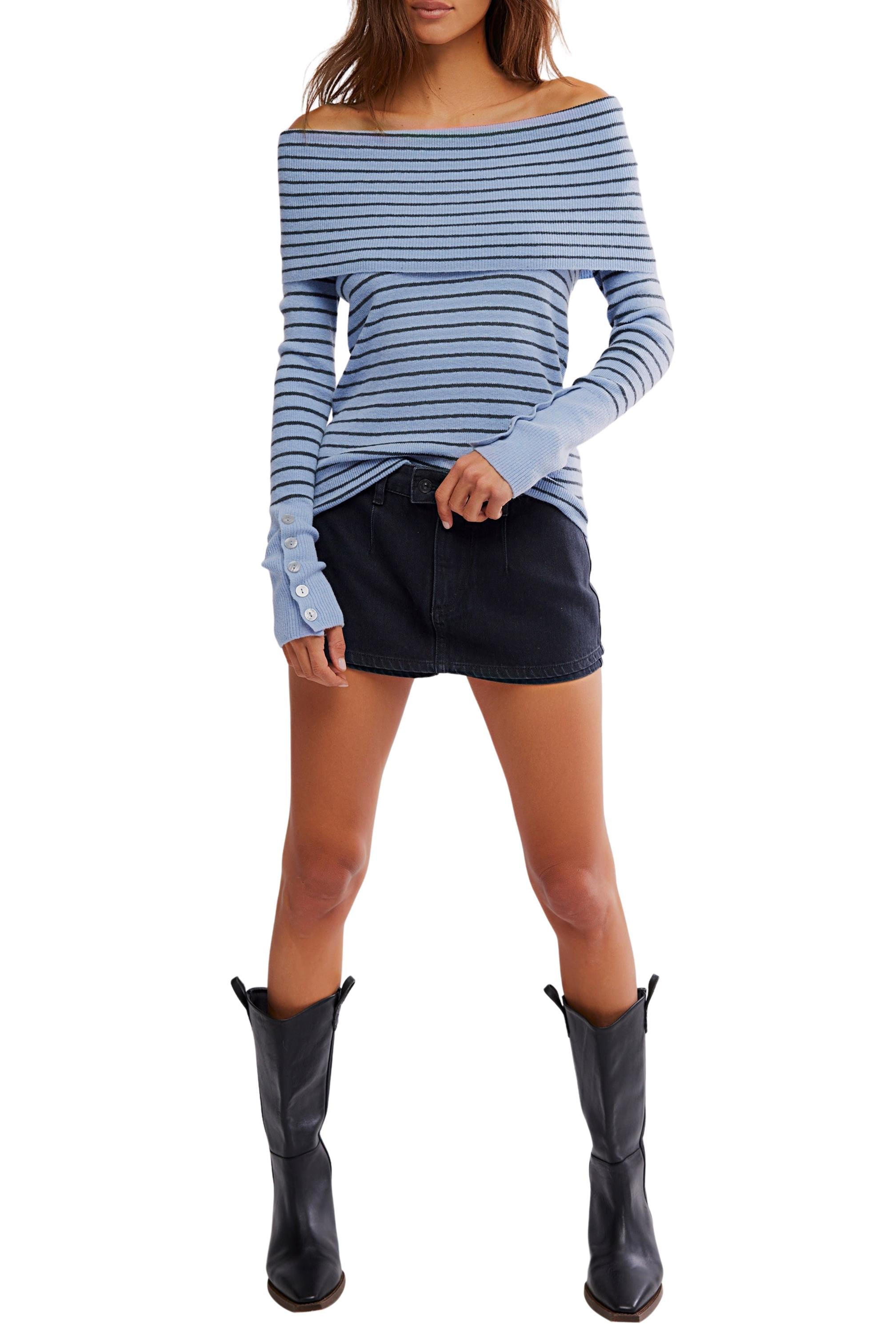 A woman models the Evergreen Sweater by Free People—a blue off-the-shoulder, striped top—paired with a black mini skirt and knee-high boots. She stands with legs apart against a white background; her head is not visible.