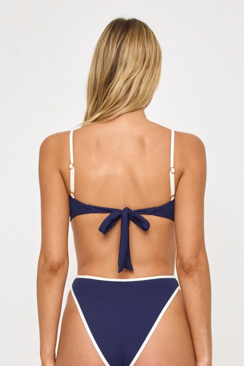 A woman with long blonde hair is shown from the back, wearing the L*Space Hazel Colorblock Bralette Top in Midnight/Cream. The ribbed bikini top features thin adjustable straps and a bow tied at the back, with navy blue fabric accented by white trim. The background is plain and light-colored.