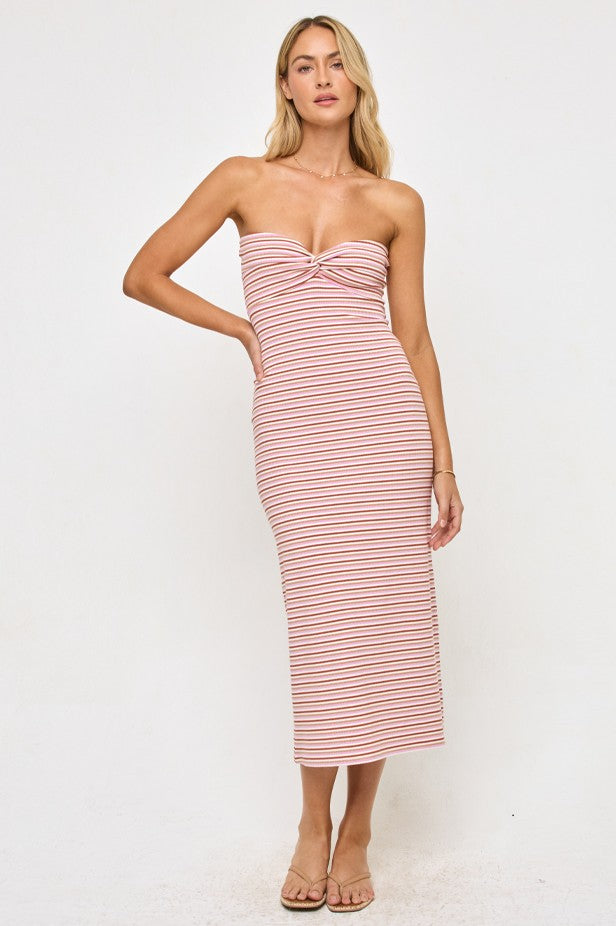 A woman stands against a white background wearing the L*Space Lisette Midi Dress in AZALEA STRIPE, a strapless style with pink and white stripes and a twist detail at the bodice. The fitted, midi-length dress is paired with nude sandals.
