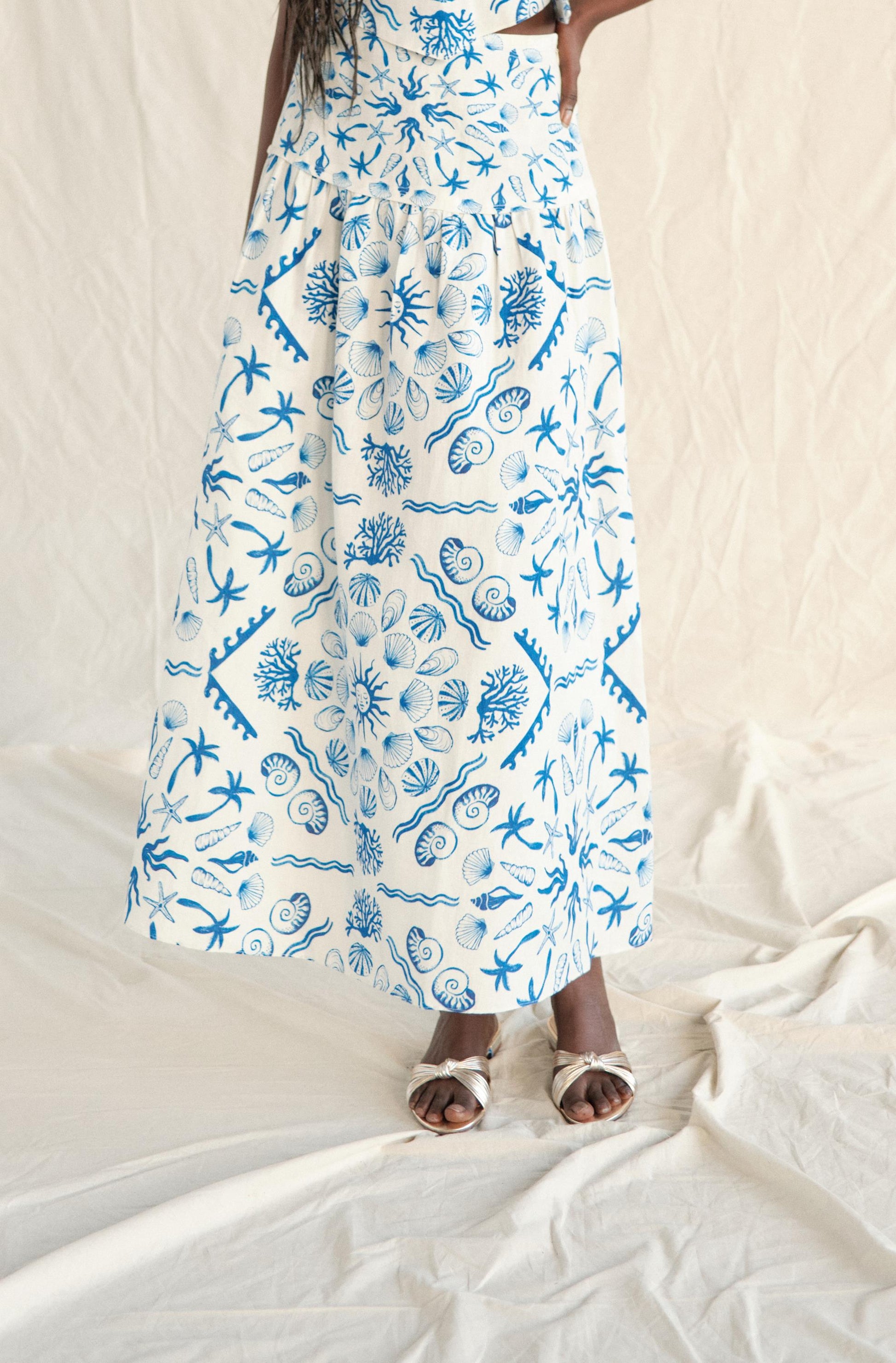 A person is seen from the waist down wearing the Sable Set Skirt by Saylor in Blue Multi, featuring blue floral and nature-inspired patterns, paired with silver sandals on a white fabric background.