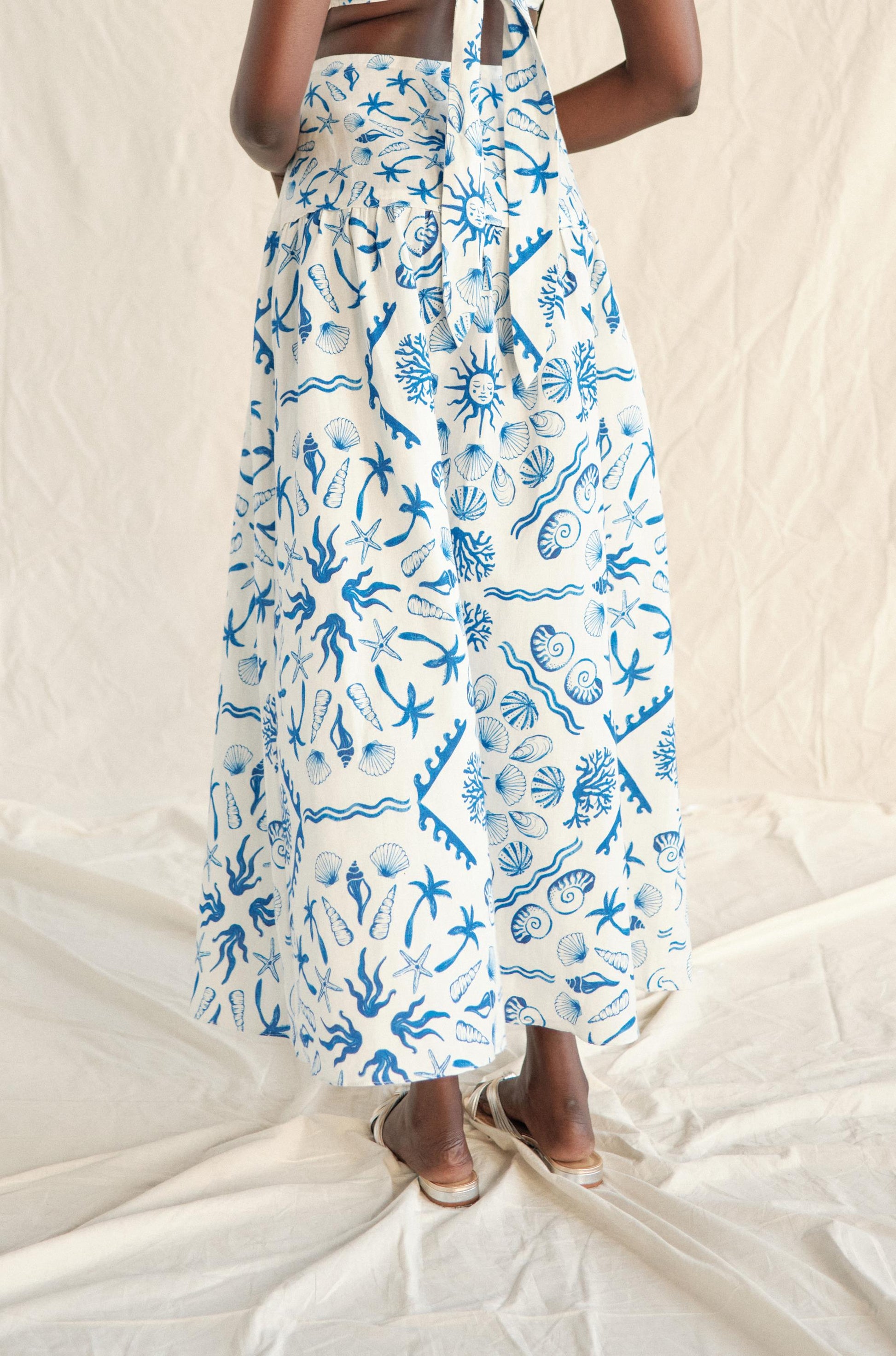 A person stands barefoot on cream fabric, wearing the Saylor Sable Set Skirt—white with blue sea-inspired patterns of shells and corals—against a light textured backdrop. The upper body is mostly out of frame.