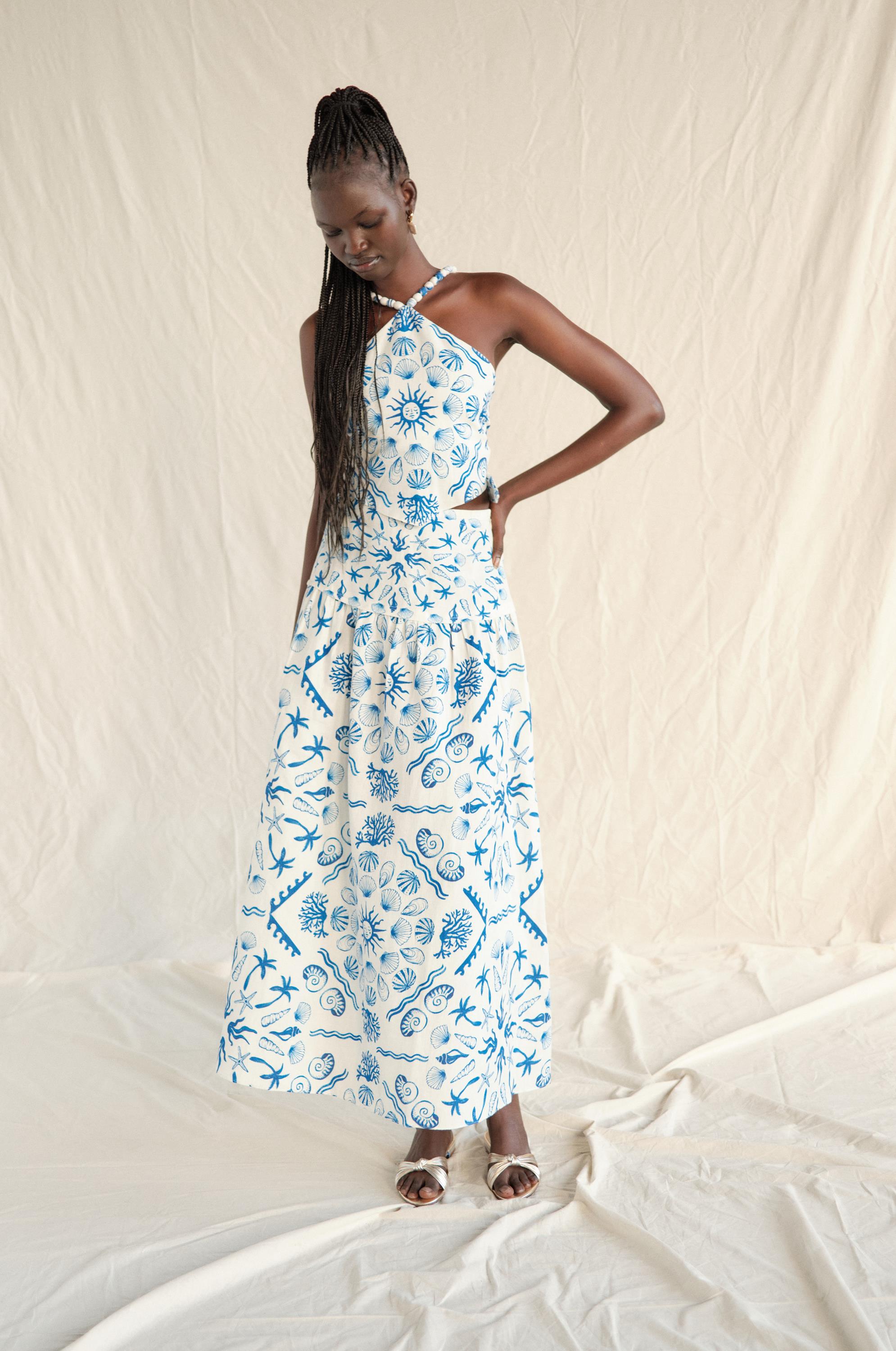 A woman wearing the Sable Set Skirt by Saylor stands against a neutral fabric backdrop, looking down with one hand on her hip. She wears sandals and has long braided hair.