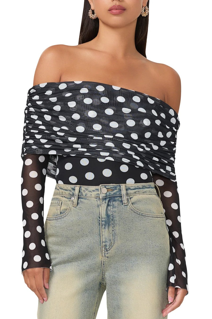 A woman wears the AFRM Maxine Bodysuit, an off-shoulder long sleeve black top with white polka dots and sheer sleeves, paired with light blue high-waisted jeans. Her head is partly out of frame.