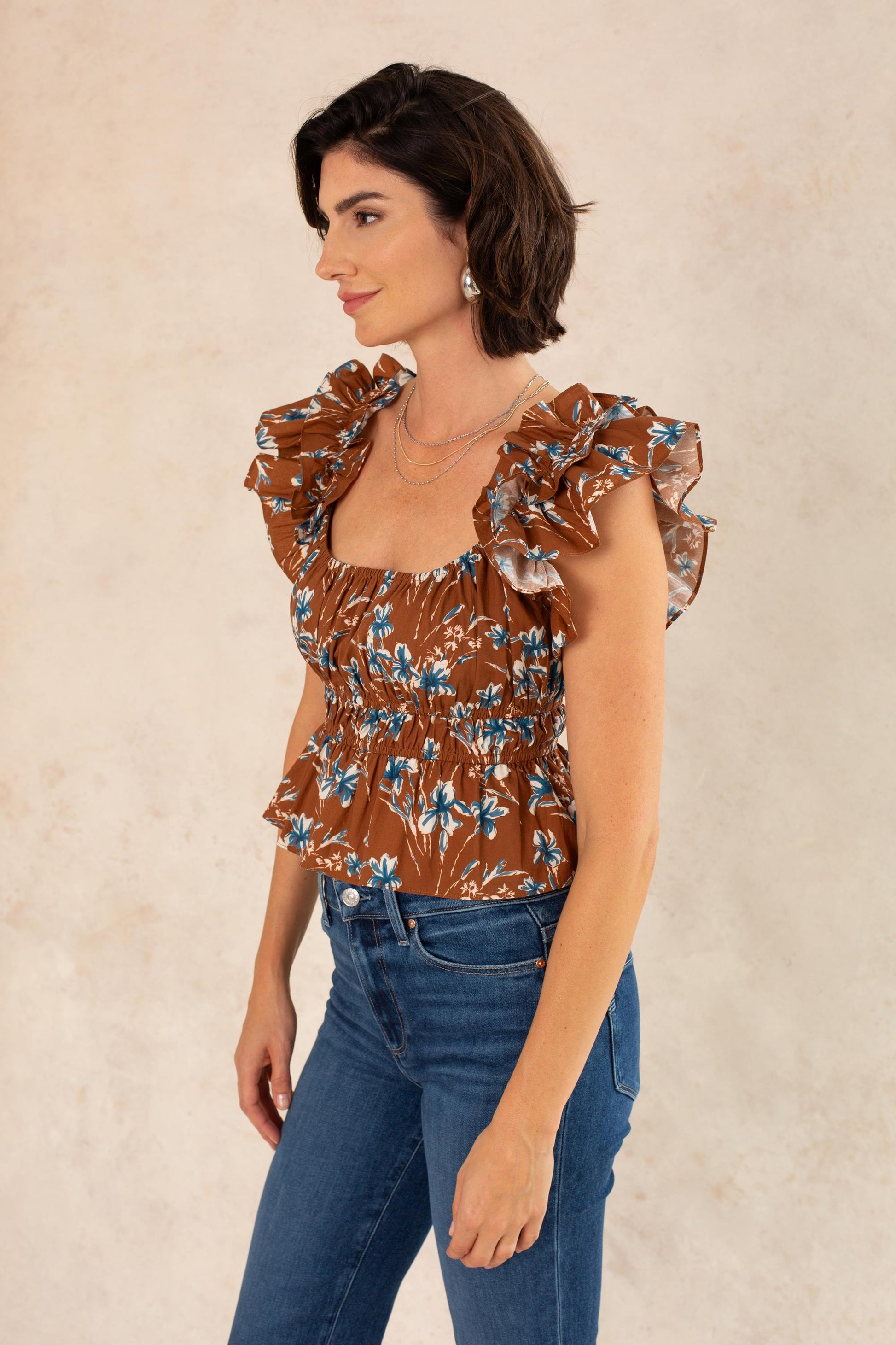 A woman with short dark hair wears the Georgie Avery Top in Brown Floral—a blouse featuring a smocked bodice—paired with blue jeans. She stands sideways against a light, neutral background, accessorized with layered necklaces and earrings, looking relaxed and confident.