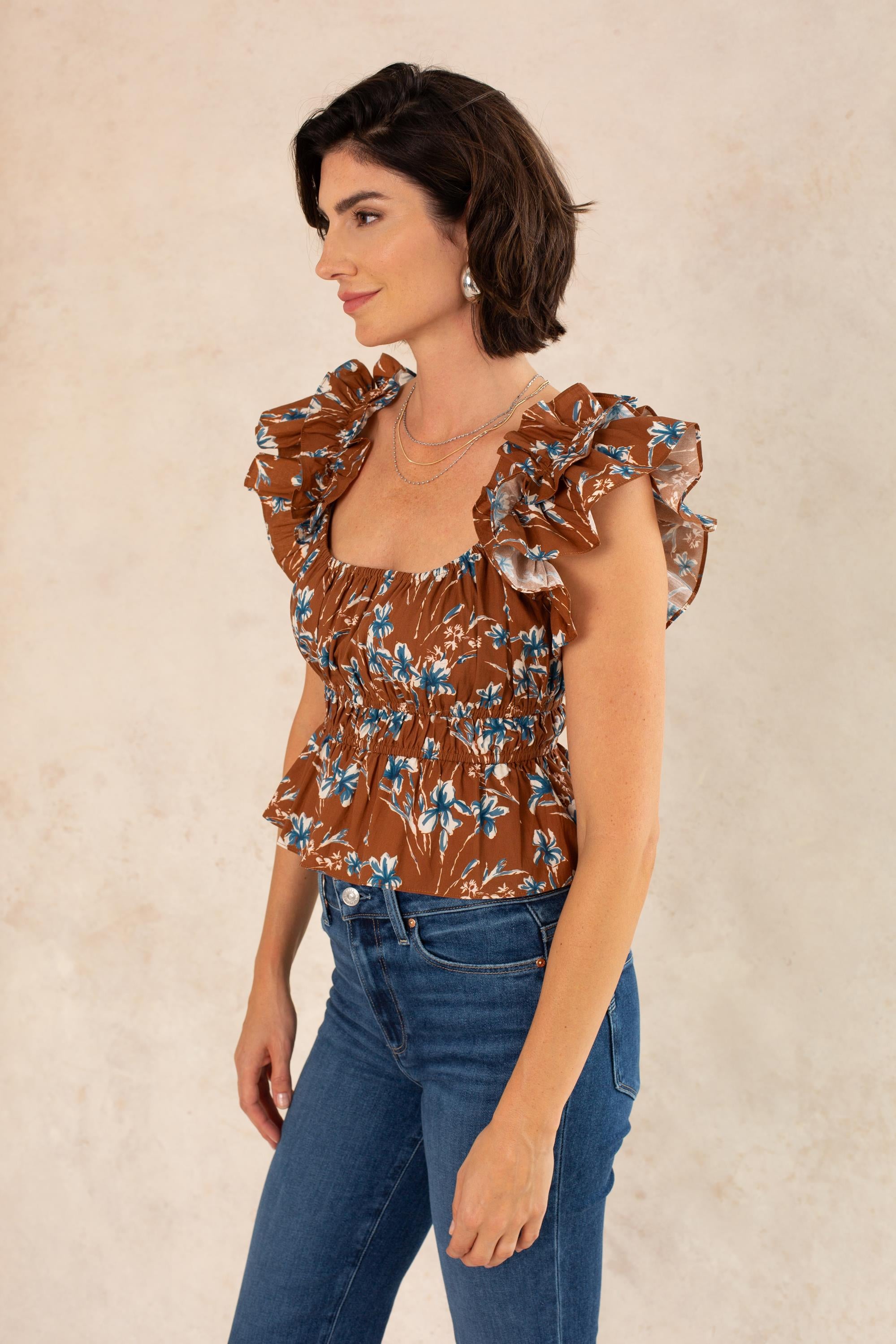 A woman with short dark hair wears the Georgie Avery Top in Brown Floral—a blouse featuring a smocked bodice—paired with blue jeans. She stands sideways against a light, neutral background, accessorized with layered necklaces and earrings, looking relaxed and confident.