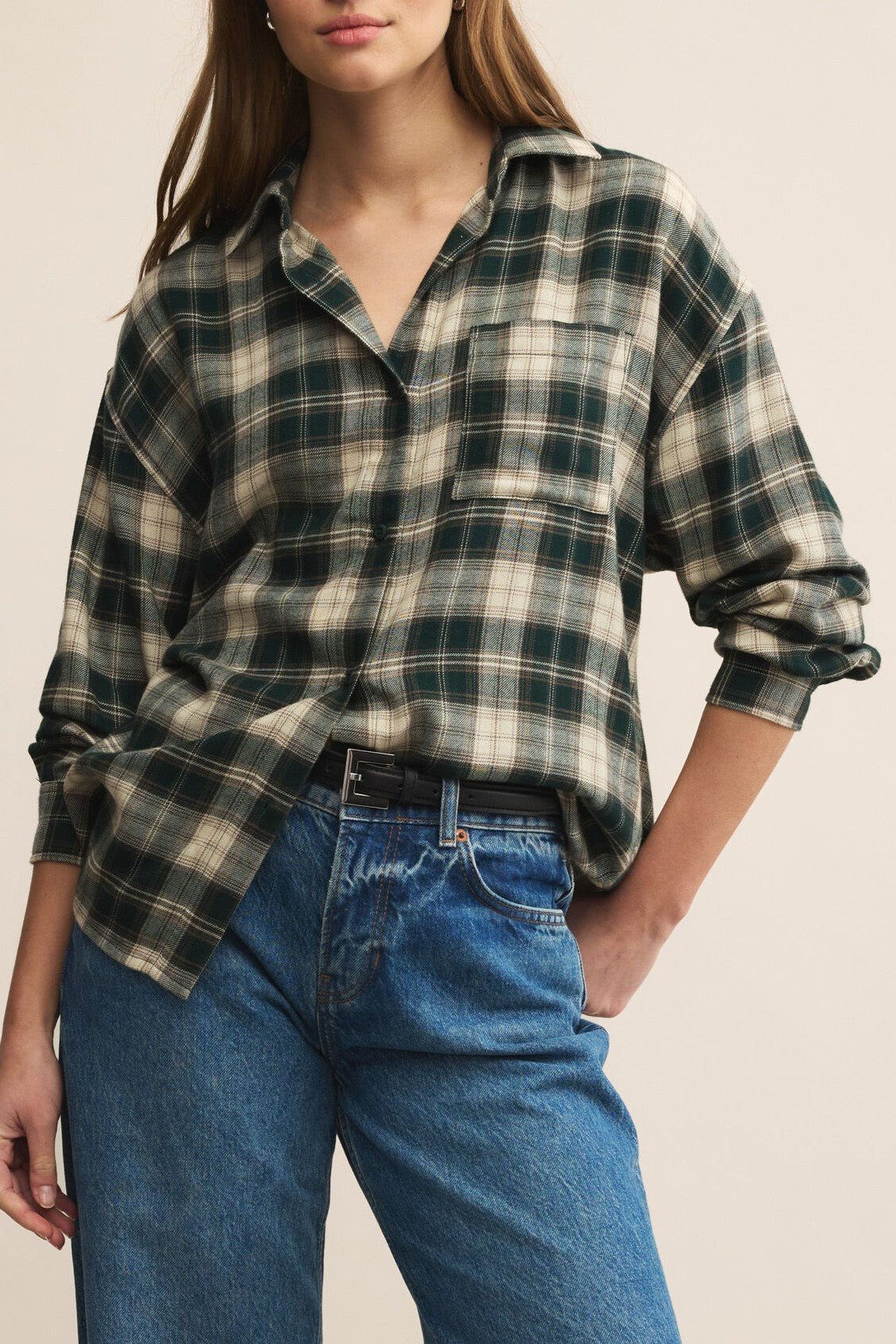 A person wearing the Z Supply River Plaid Button Up in Garland Green with blue jeans stands with one hand in their pocket against a plain background.