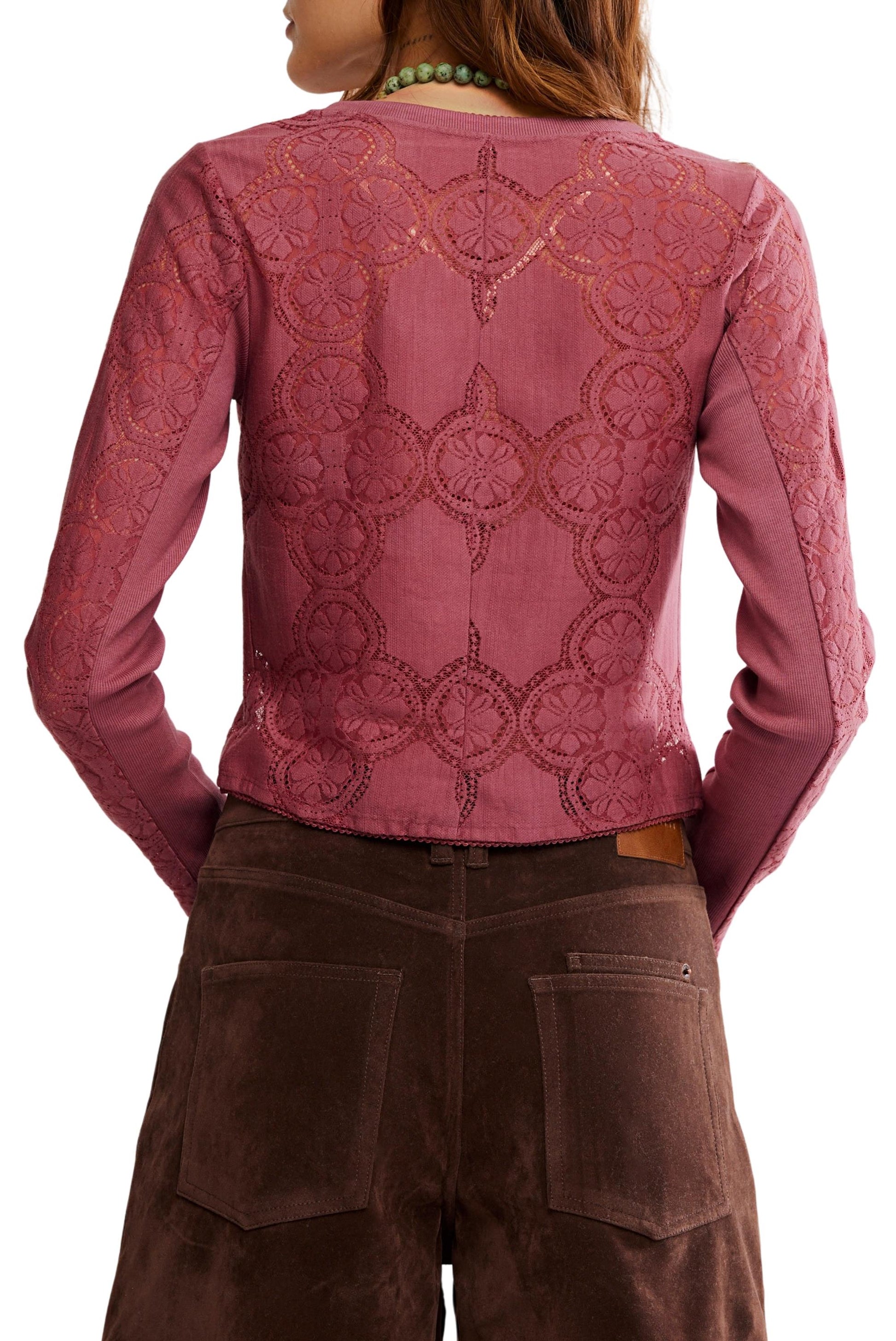 A woman is shown from the back wearing the Free People Jae Knit Long Sleeve—a sheer, pink cotton-lace top with a circular lace pattern, solid pink center panel, and elegant button-front detailing—paired with brown suede pants.