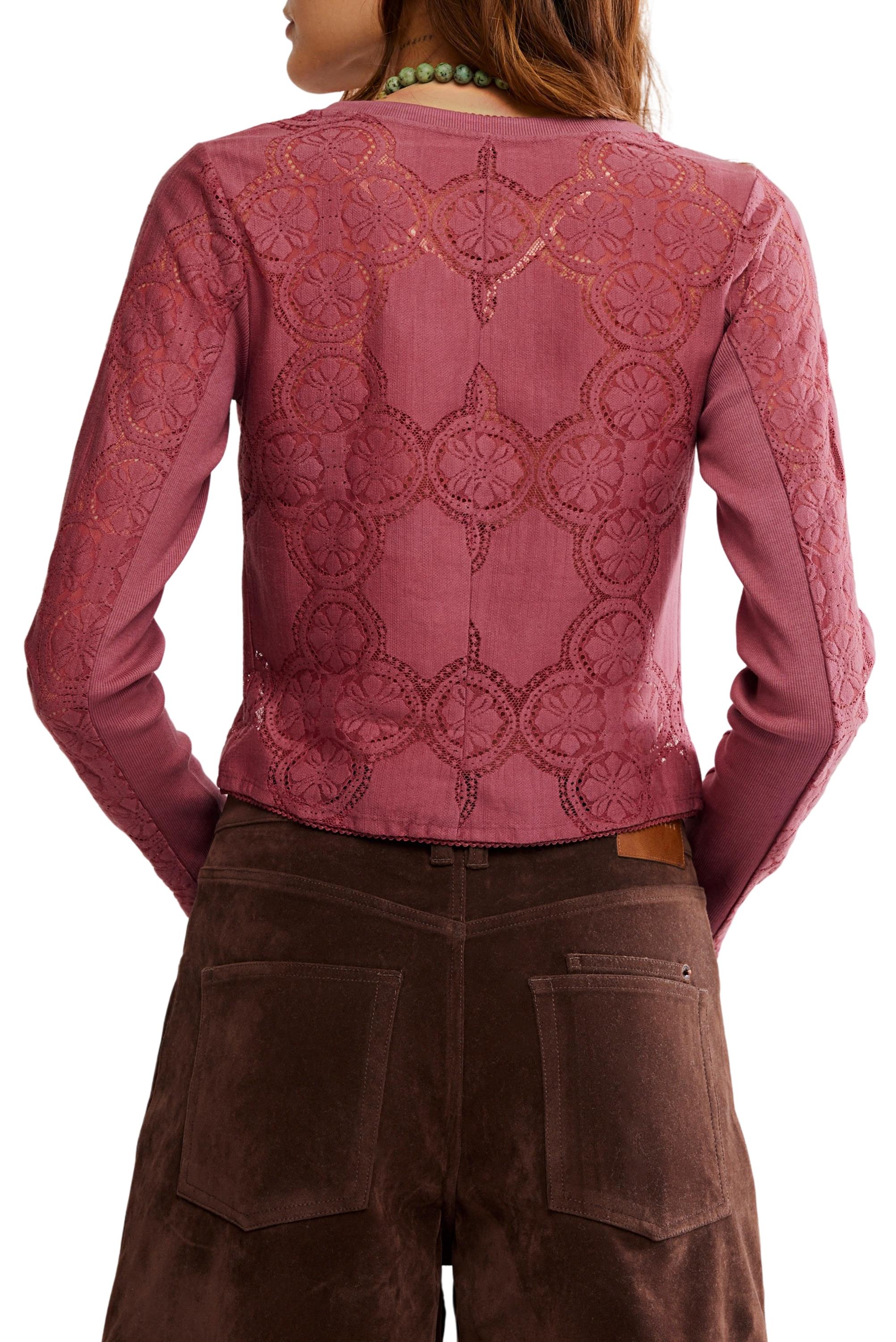 A woman is shown from the back wearing the Free People Jae Knit Long Sleeve—a sheer, pink cotton-lace top with a circular lace pattern, solid pink center panel, and elegant button-front detailing—paired with brown suede pants.
