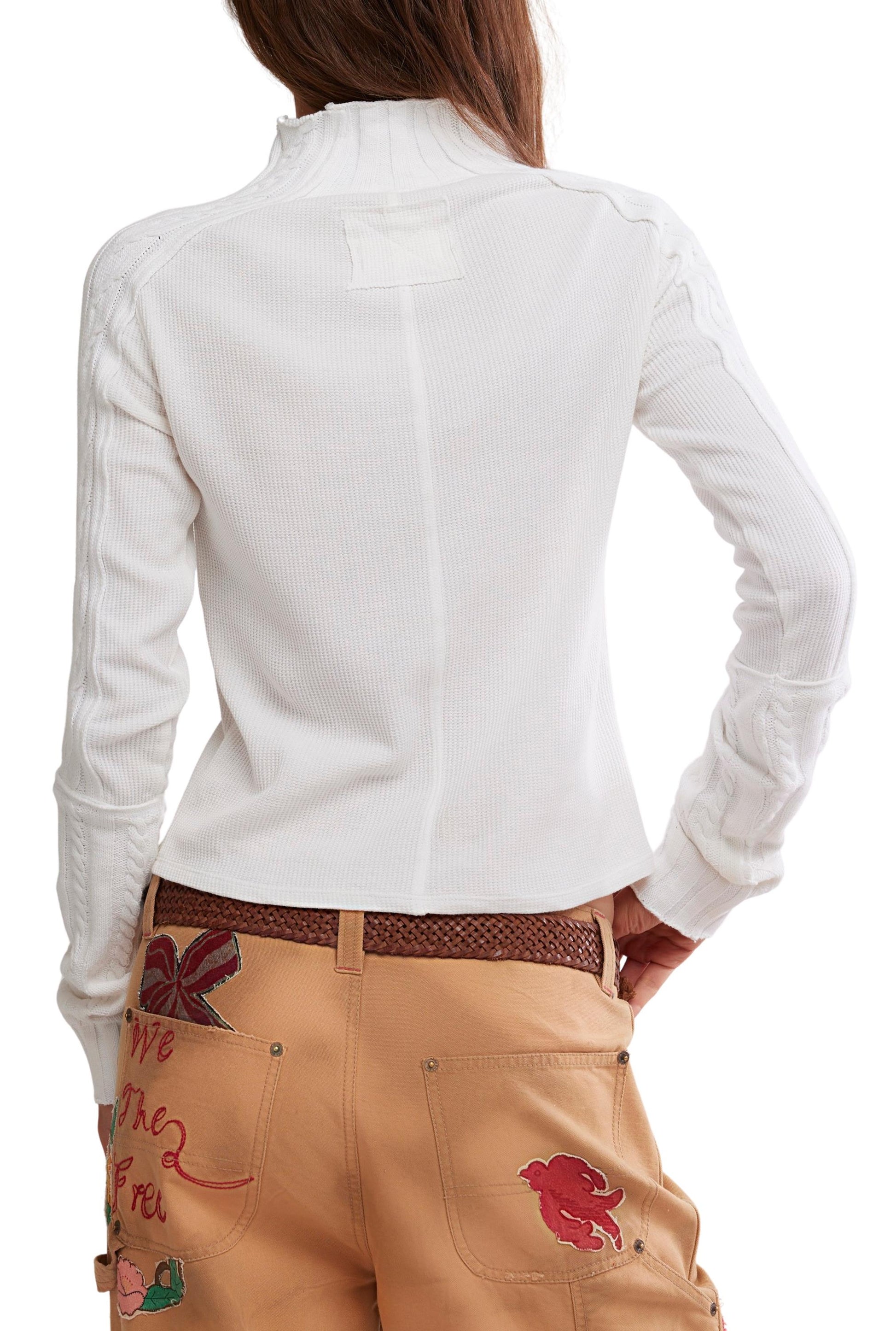 A person wearing the Free People Wtf Breezy Swit, a white, long-sleeve, semi-fitted cotton knit top with a subtle ribbed texture, paired with tan pants featuring colorful embroidered designs stands facing away, one hand on their hip.