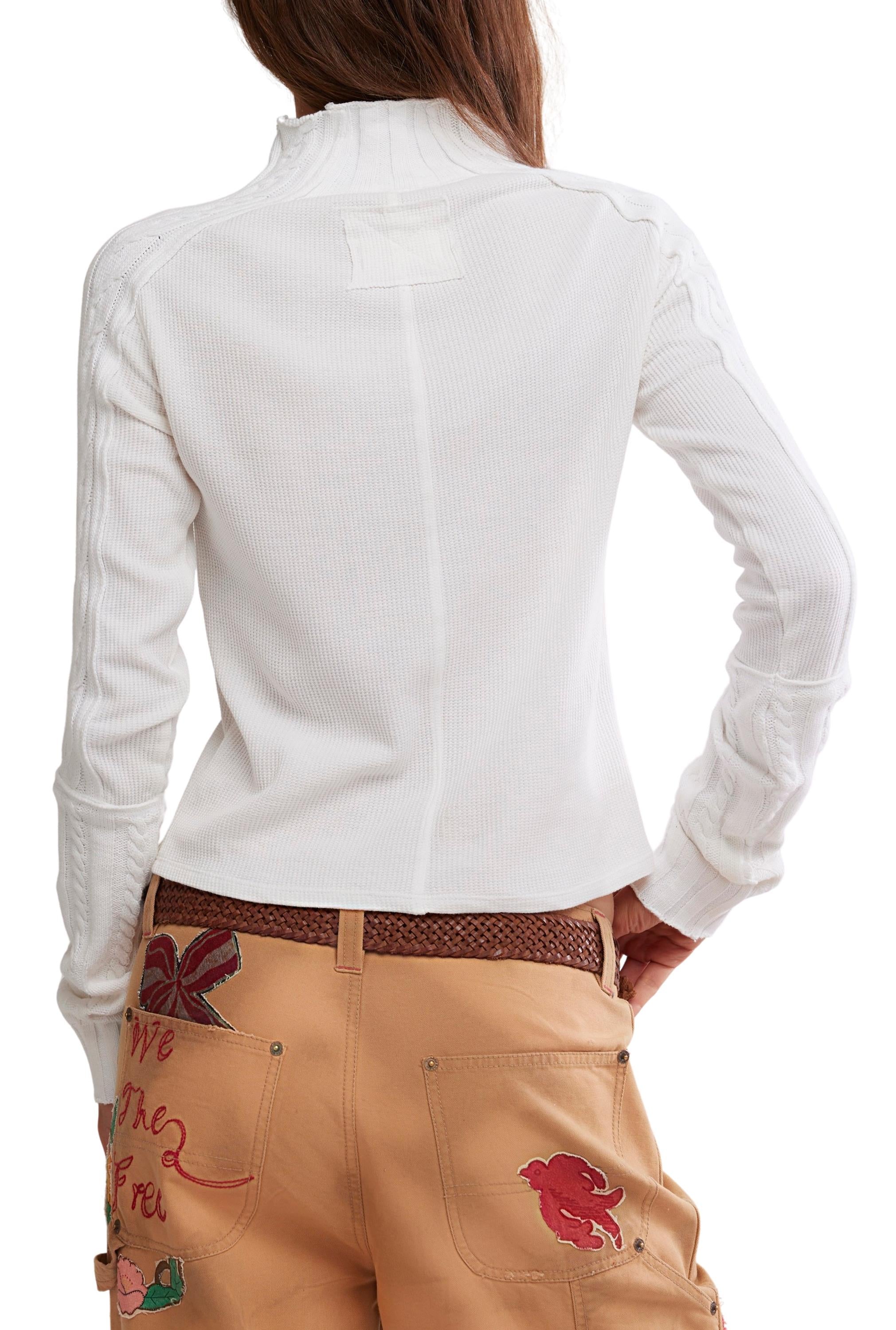 A person wearing the Free People Wtf Breezy Swit, a white, long-sleeve, semi-fitted cotton knit top with a subtle ribbed texture, paired with tan pants featuring colorful embroidered designs stands facing away, one hand on their hip.