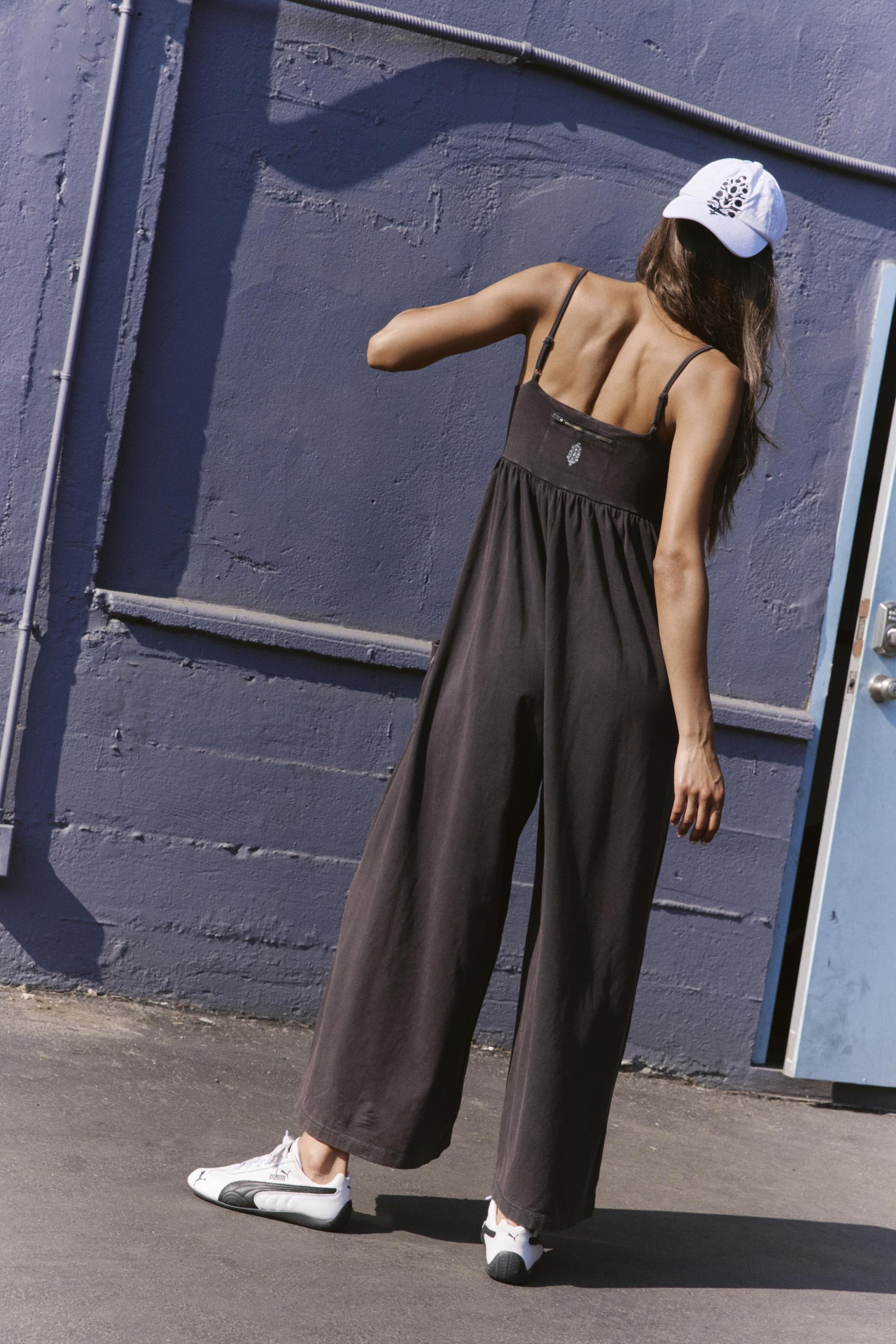 A woman stands with her back to the camera, wearing the FP Movement Hot Shot Strappy Jumpsuit in Black, paired with white sneakers and a white baseball cap. She is outside by a blue wall and door, sunlight casting distinct shadows.