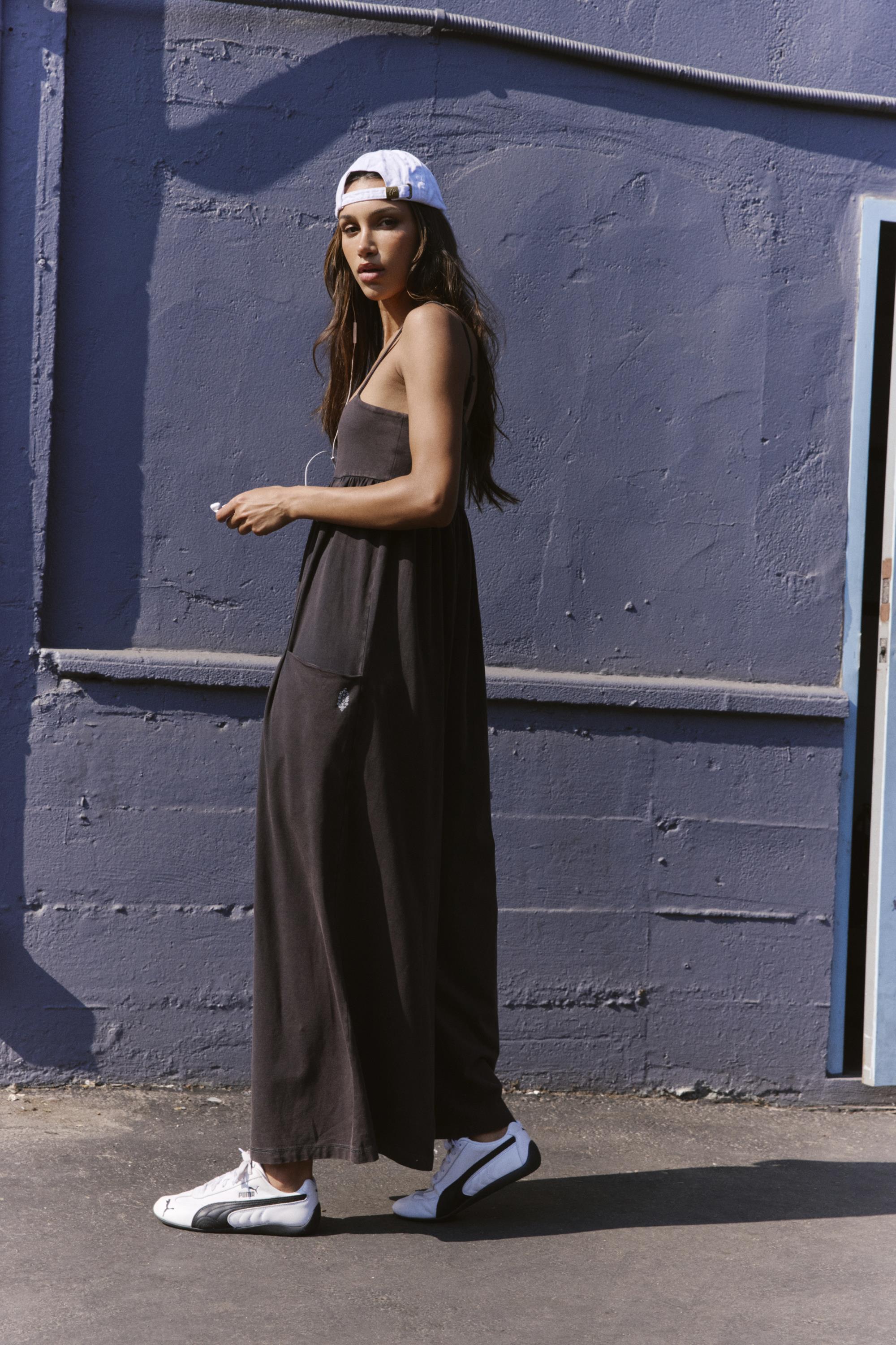 A woman in the FP Movement Hot Shot Strappy Jumpsuit in Black, white sneakers, and a backward white cap walks outside by a purple wall, glancing over her shoulder at the camera.