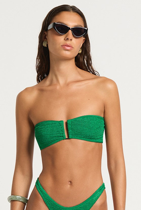 A woman wearing the Bond-Eye Emerald Shimmer Blake Bandeau in EMERALD SHIMMER and black cat-eye sunglasses poses against a plain background. She has long brown hair and accessorizes with hoop earrings and a chunky bracelet.