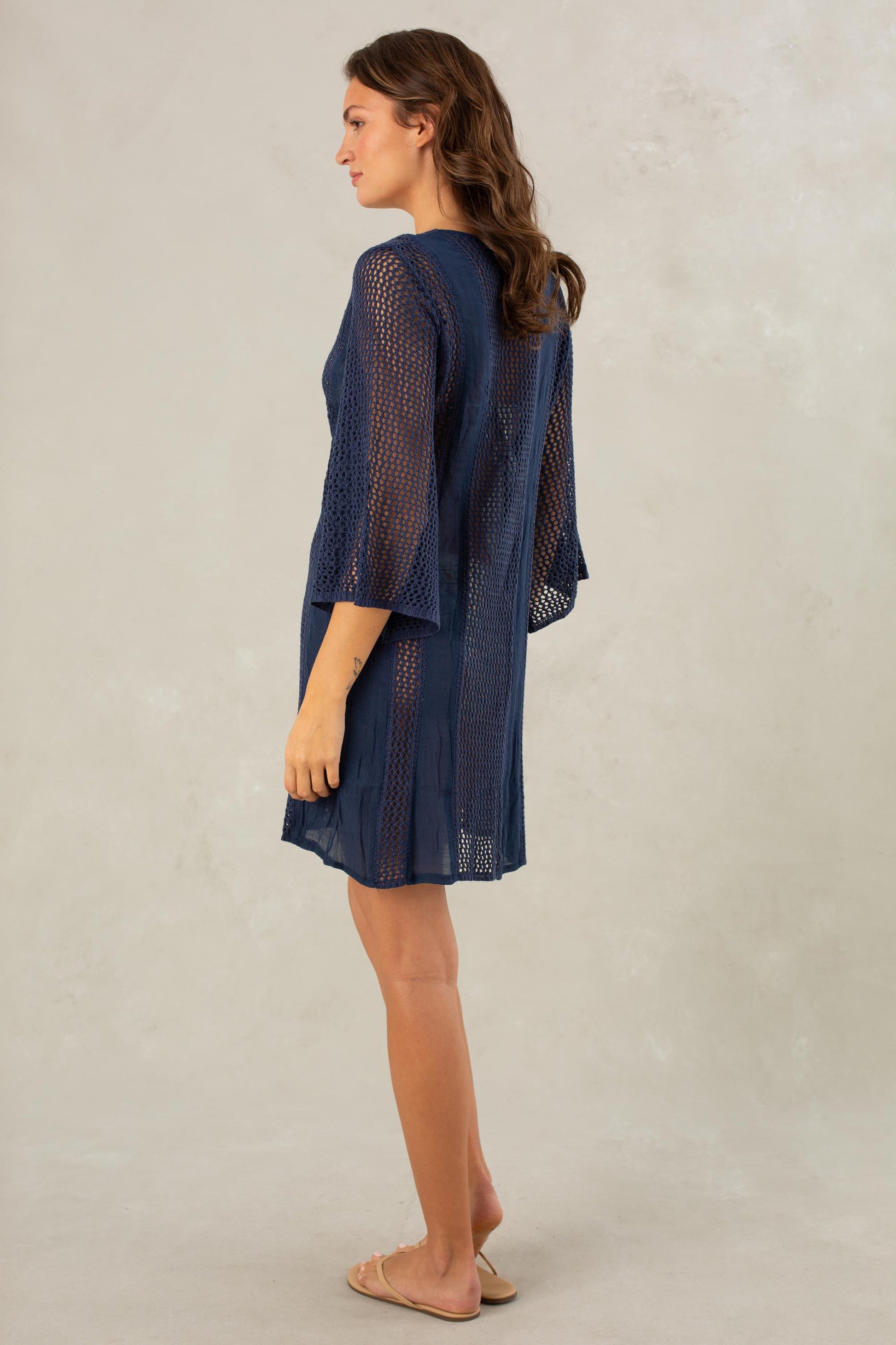 A woman stands sideways wearing the Isla Adriana Cover Up Kimono Coverup in navy blue—a knee-length, sheer dress with three-quarter sleeves and delicate crochet detailing along the center front—paired with tan sandals against a light gray background.