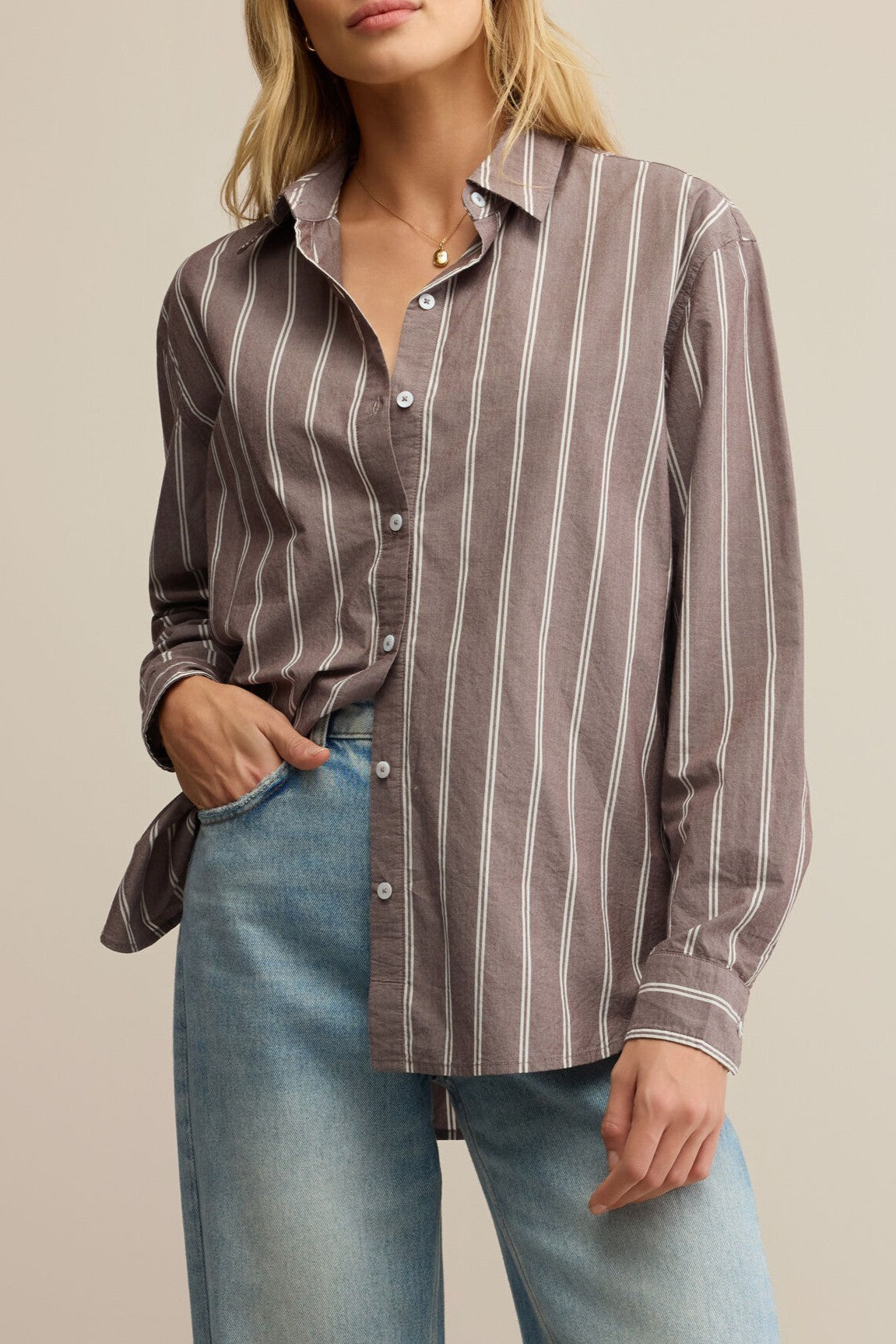 A woman stands against a plain beige background wearing the Z Supply Seaport Striped Poplin Shirt, featuring a relaxed fit and brown stripes, paired with light blue jeans and a simple gold necklace.