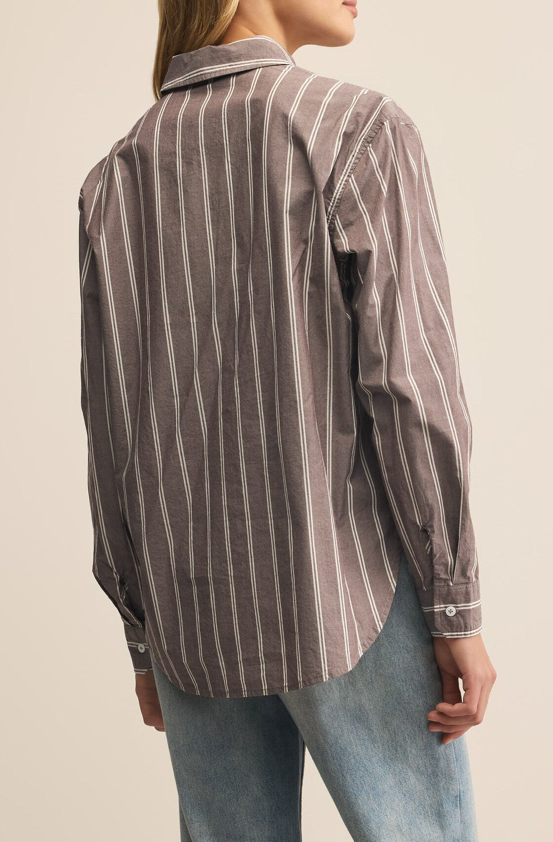 A person is seen from the back against a beige background, wearing Z Supply's Seaport Striped Poplin Shirt in brown and white with long sleeves, paired with light blue jeans.