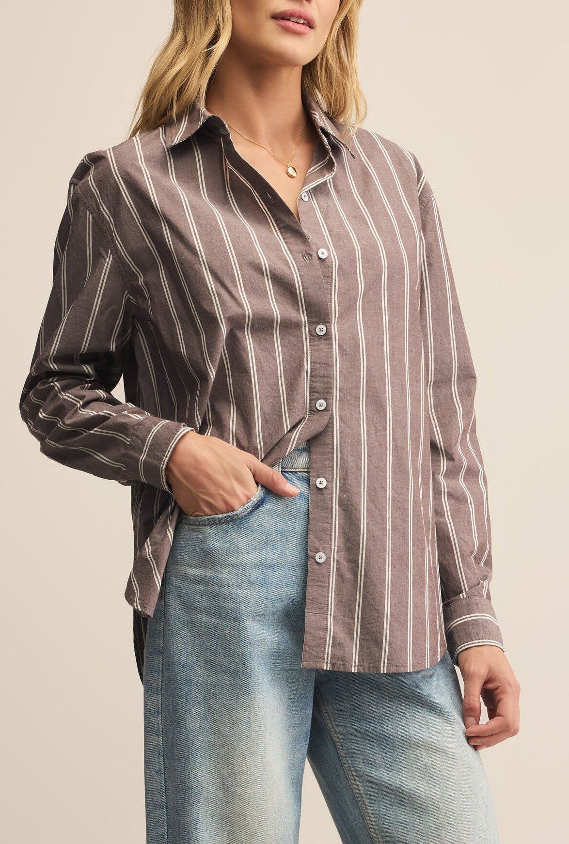 A woman wears the Z Supply Seaport Striped Poplin Shirt in brown stripe with a relaxed fit and cotton fabric, paired with light blue jeans. She stands with one hand in her pocket, facing slightly to the side.