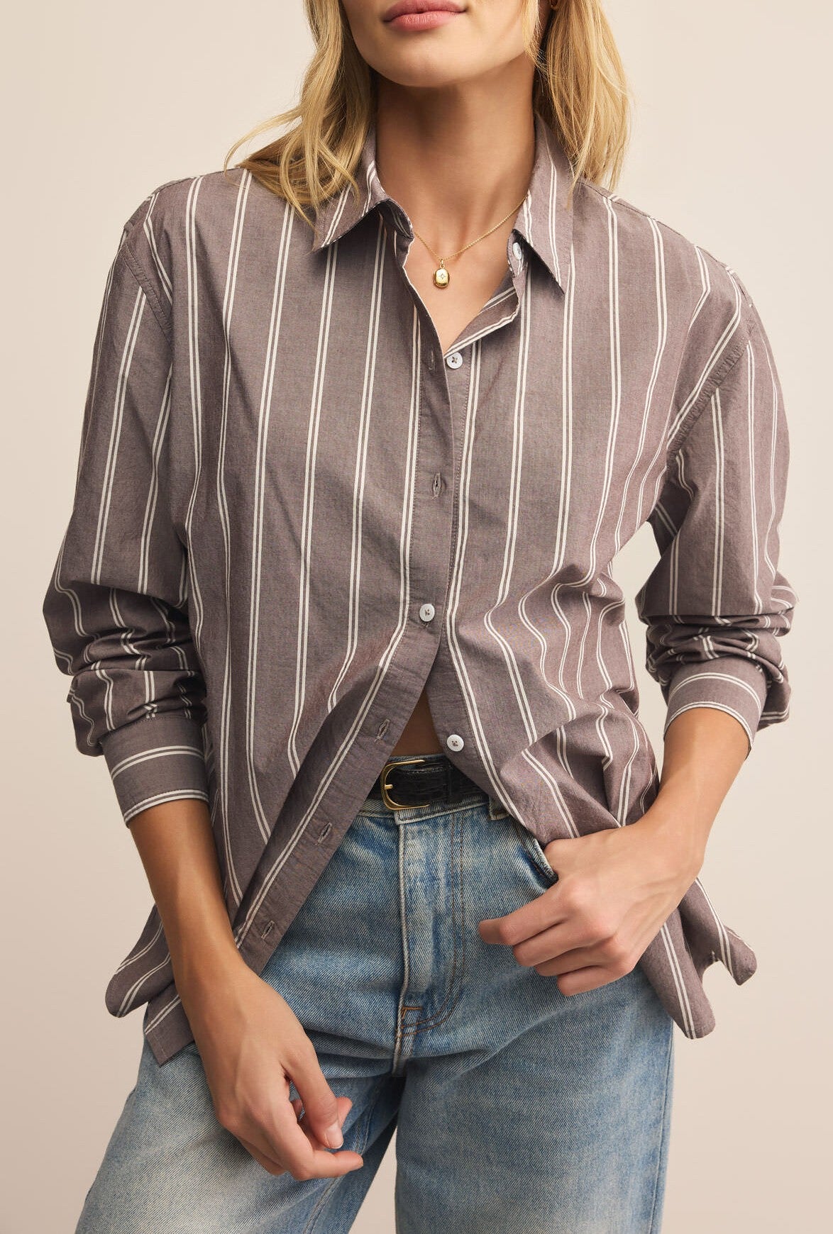 A woman wears the Z Supply Seaport Striped Poplin Shirt with rolled sleeves and light blue jeans. The shirt is partially unbuttoned and tucked in, paired with a gold pendant necklace. Her face is partially out of frame.