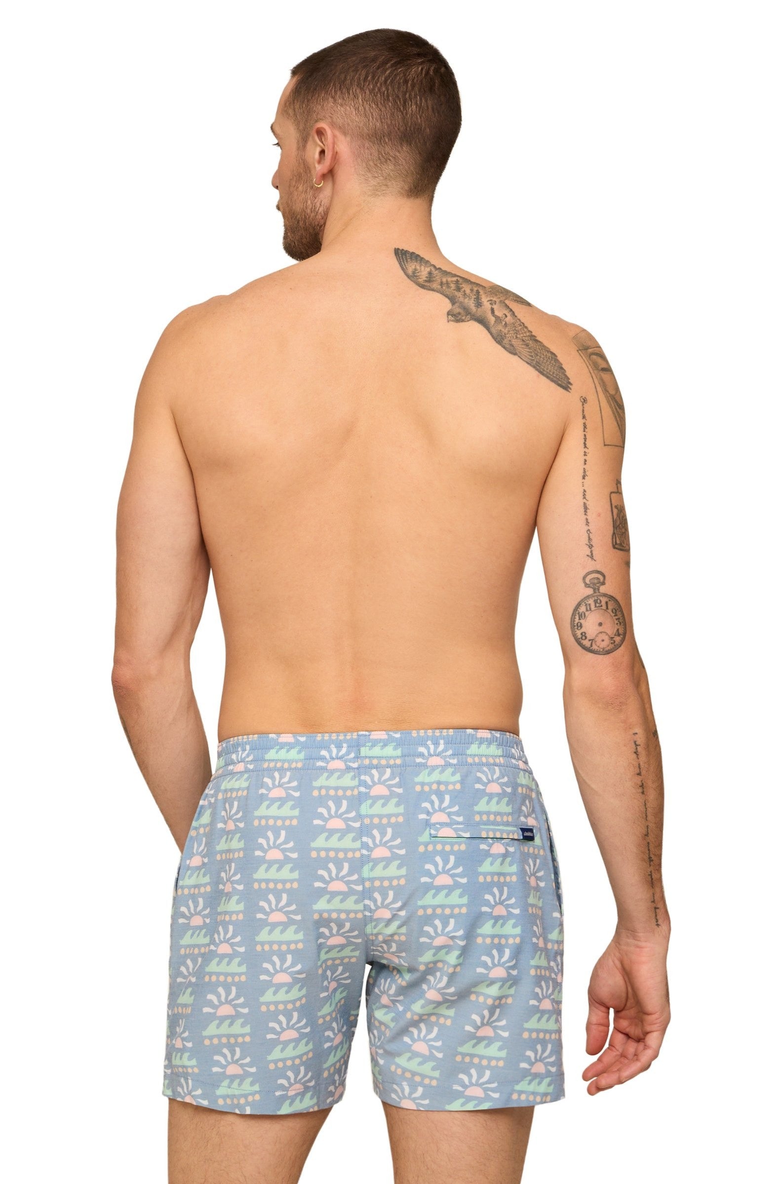 A man shown from behind wears Chubbies The Easygoers 5.5 Inch Lined Swim trunks in light blue with a pastel sun and wave print, shirtless with visible tattoos on his back and arm. Back pocket is visible on the right side.