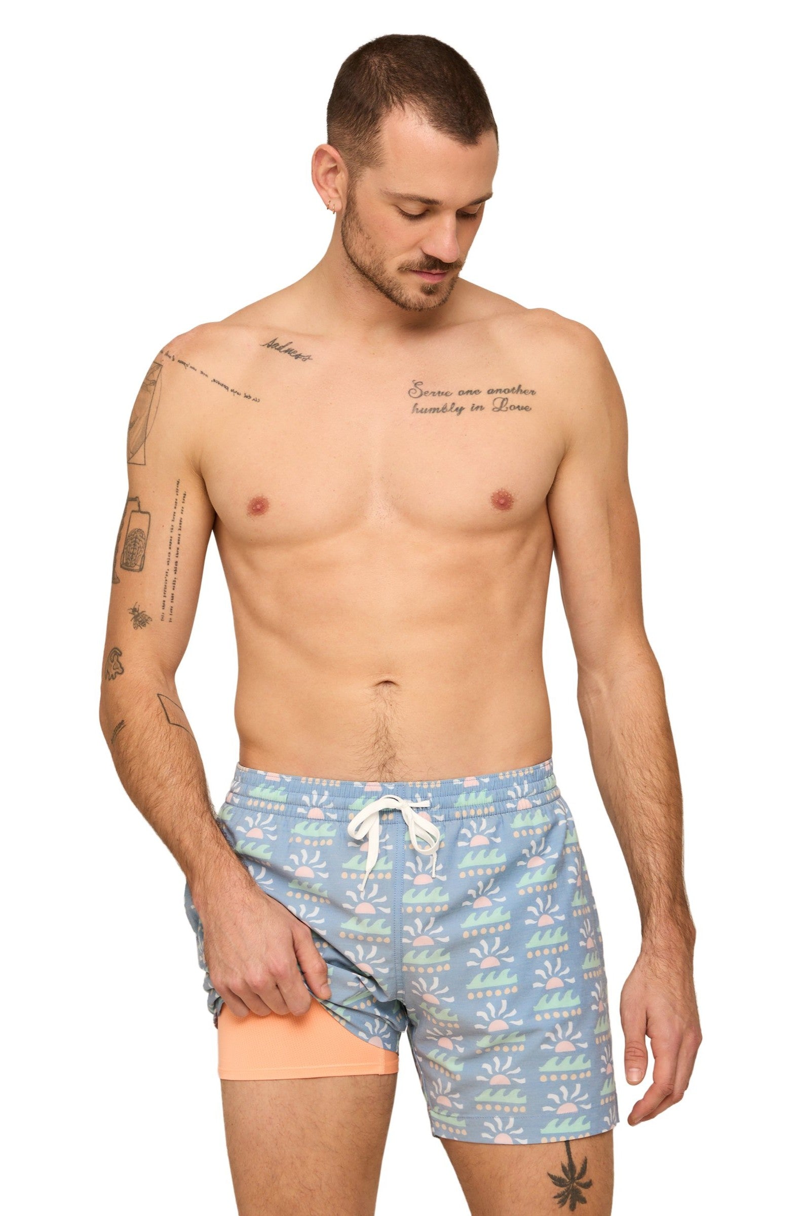 A man with short hair and tattoos is wearing Chubbies’ The Easygoers 5.5 Inch Lined Swim trunks, featuring a light blue pattern and white drawstring, and revealing peach compression liner as he pulls up one leg.