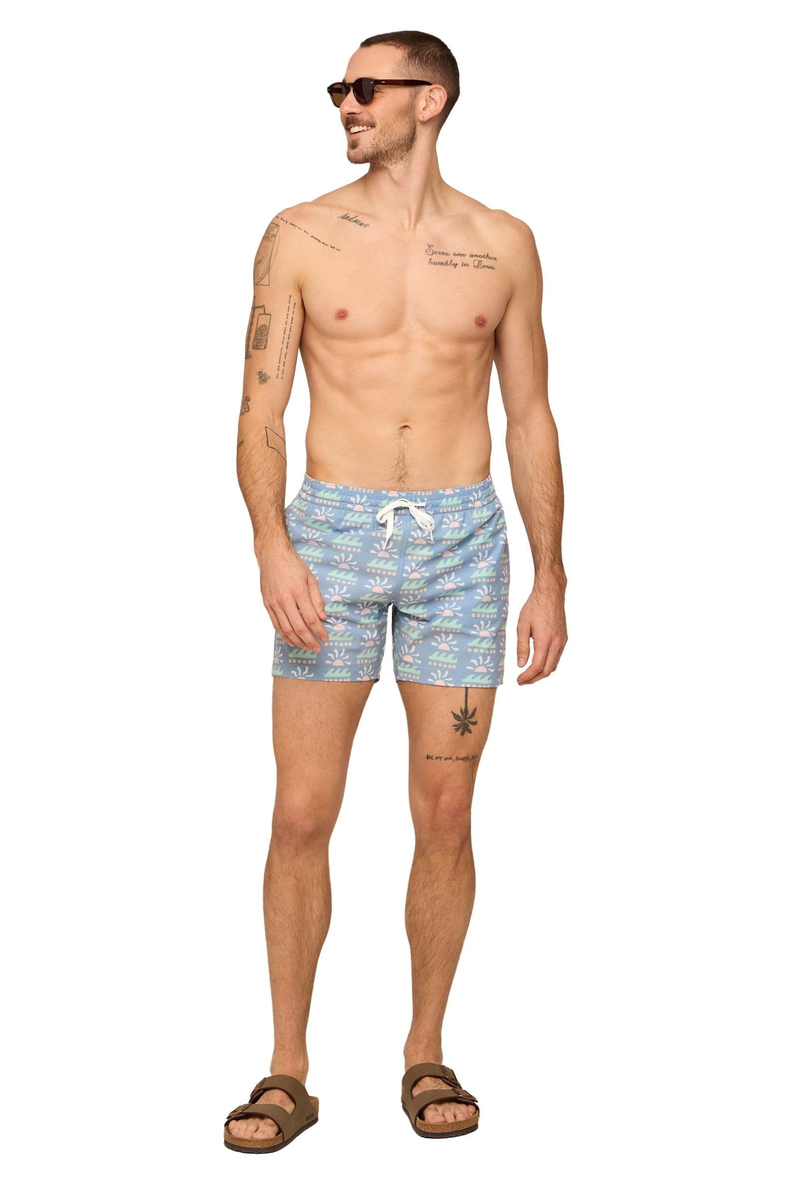 A man in Chubbies' The Easygoers 5.5 Inch Lined Swim trunks with a sun and wave pattern, sunglasses, a chest tattoo, and brown sandals stands barefoot.