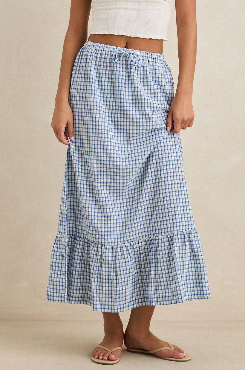 A person wears the Rhythm Lola Check Tiered Maxi Skirt, a blue-and-white checkered, high-waisted skirt with a ruffled hem, paired with a white crop top and beige flip-flops, standing against a neutral background.