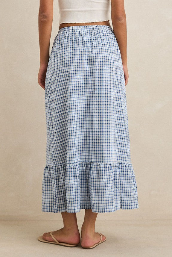 A person wearing the Rhythm Lola Check Tiered Maxi Skirt, a white top, and beige sandals stands against a plain light-colored background. The photo is taken from behind.