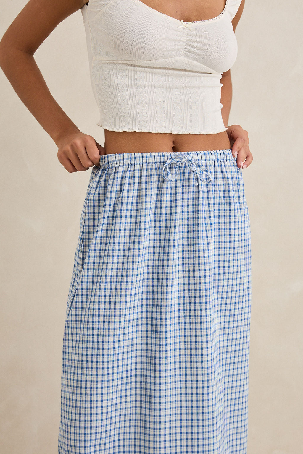 A person wears the Rhythm Lola Check Tiered Maxi Skirt, a blue and white checkered high-waisted skirt with an elastic waistband, paired with a sleeveless white crop top, standing against a neutral background.