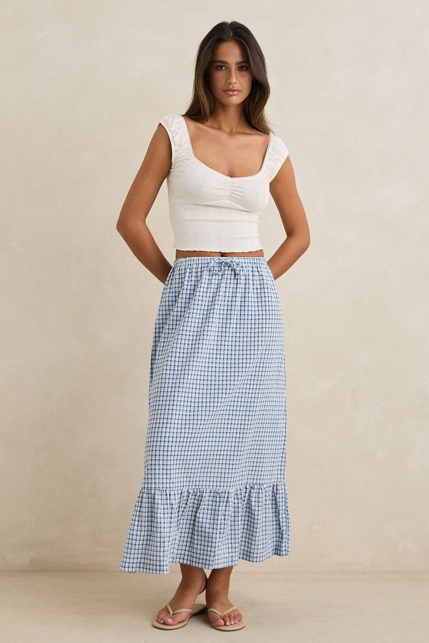 A woman with long brown hair and a relaxed expression wears the Rhythm Lola Check Tiered Maxi Skirt in blue and white gingham, a cream cap sleeve top, and beige sandals against a neutral background.