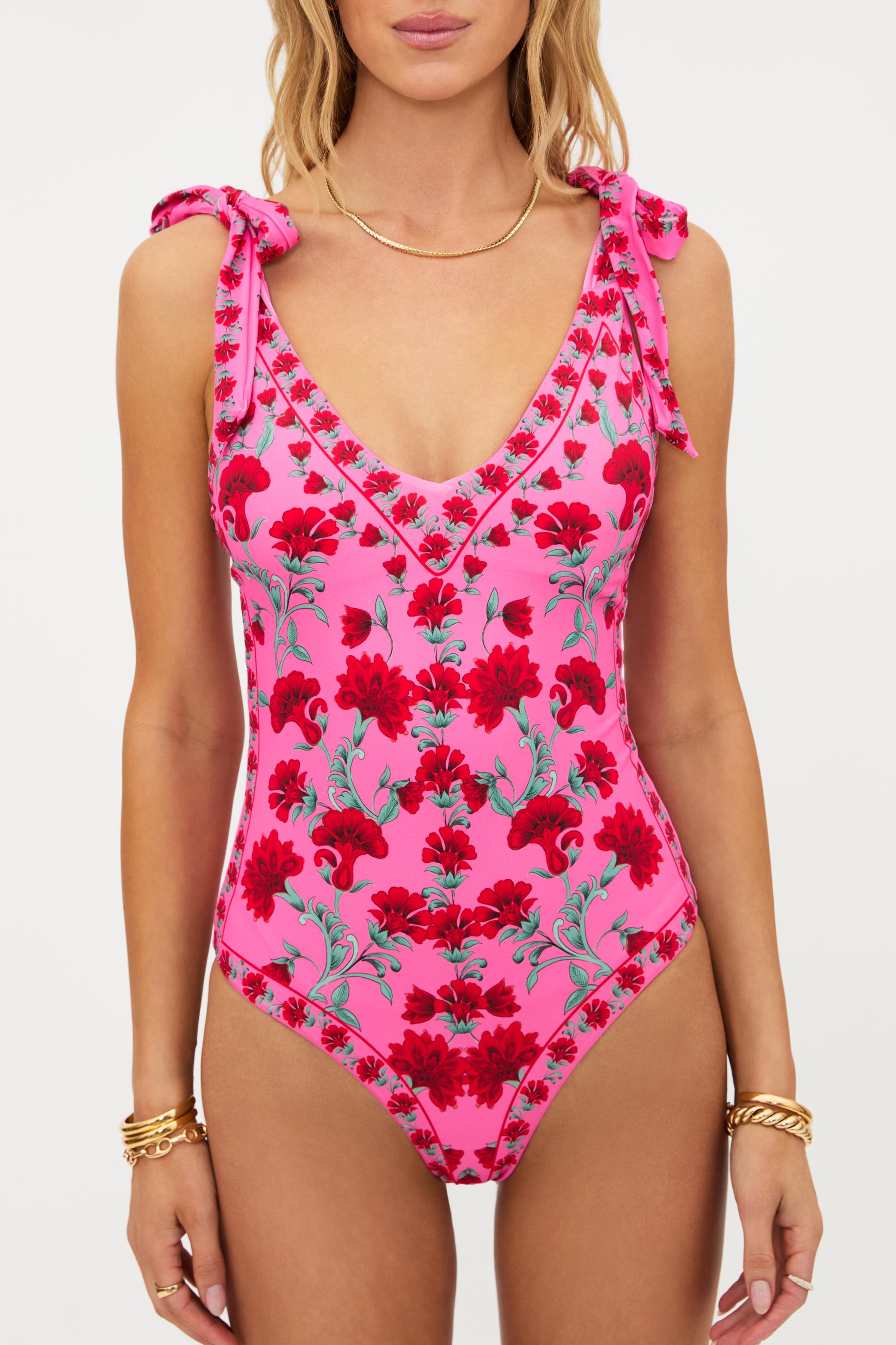 A woman wearing the Beach Riot Redwood Floral Akira One Piece swimsuit, featuring pink tie shoulder straps and a red floral pattern. This moderate coverage swimsuit showcases a V-neckline, decorative edging, and is crafted from soft nylon-spandex fabric. She completes her look with gold necklaces and bracelets.