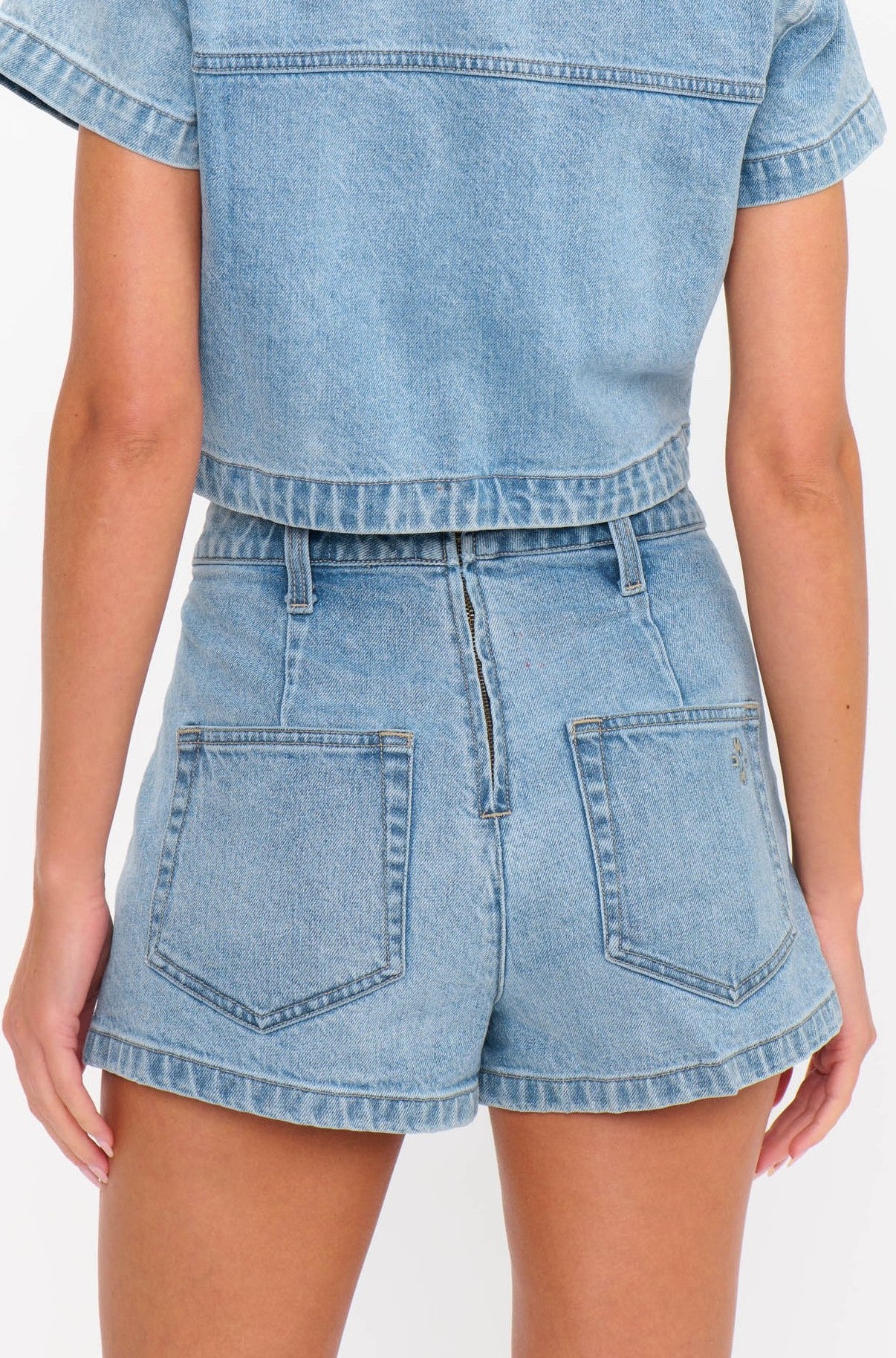 A person is shown from the back wearing the Monroe Skort by Show Me Your Mumu, a light blue stretch denim skort with two back pockets and a slightly faded wash. Their arms are relaxed at their sides, and they are paired with a matching denim top.