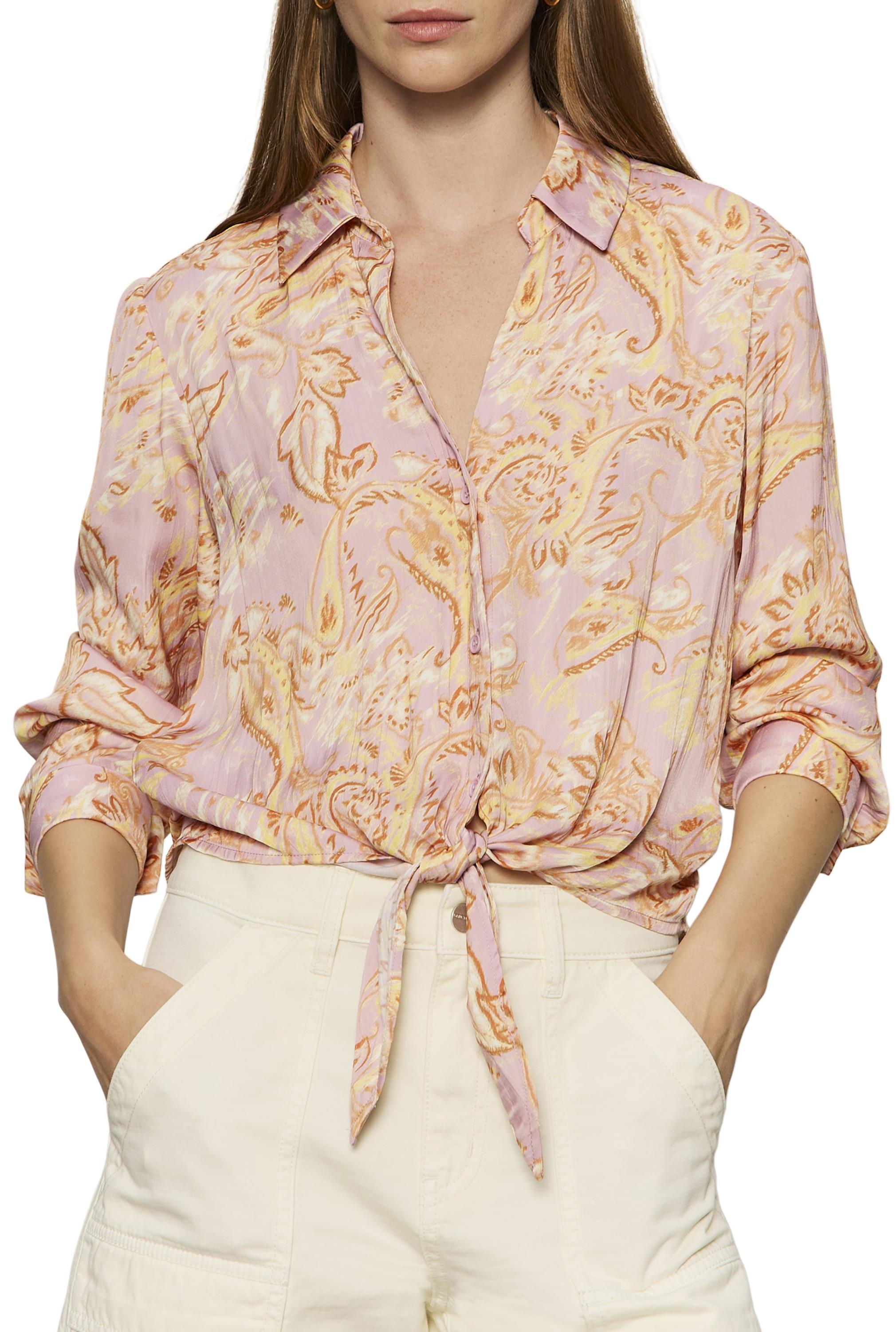 A woman wears the Sanctuary Lover Tie Shirt in WHIMSY, a light pink paisley print button-up blouse featuring an adjustable front-tie hem. She pairs it with high-waisted off-white pants, has her sleeves rolled up, and her hair is down.