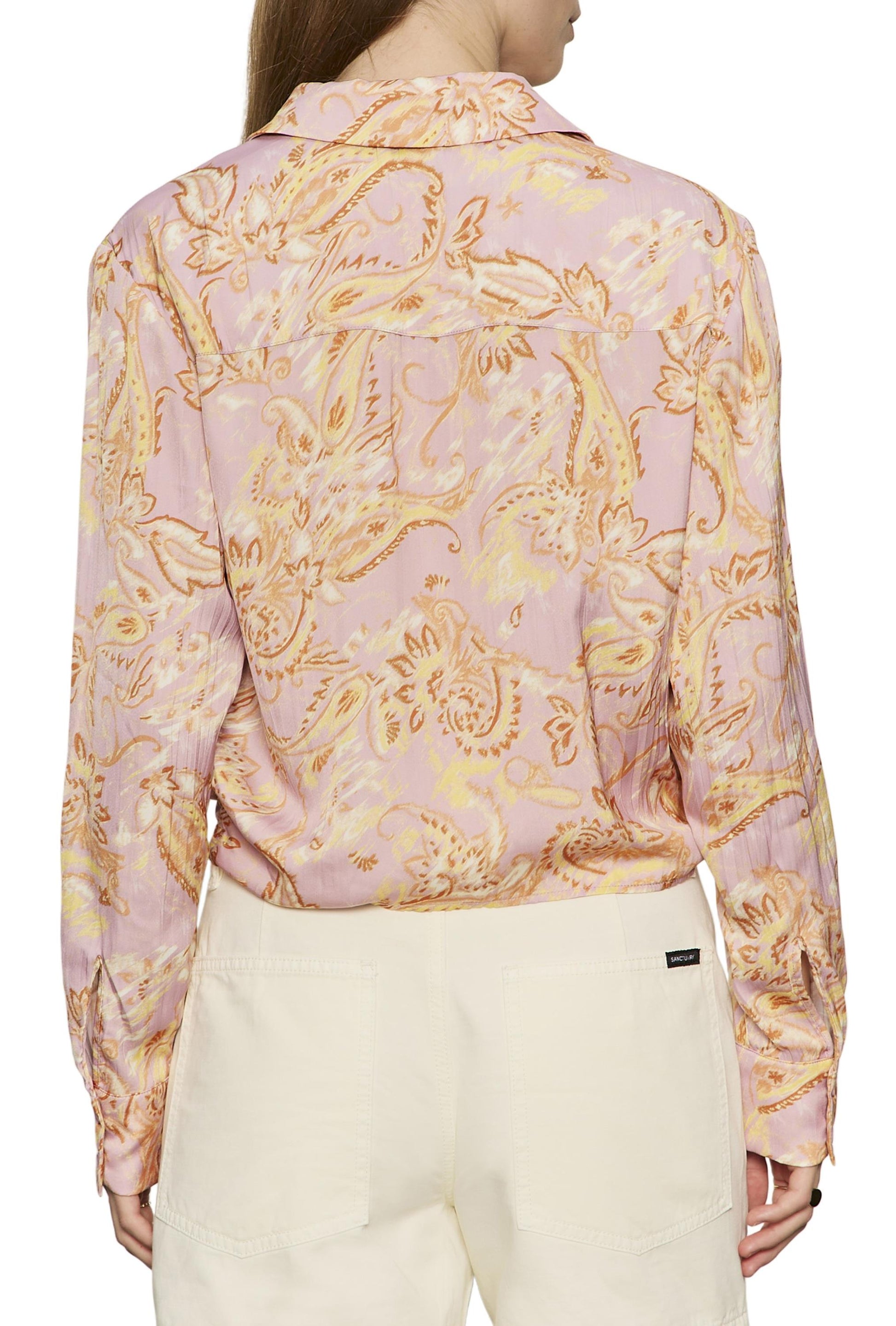 A person is photographed from the back wearing the Sanctuary Lover Tie Shirt—a long-sleeve, light pink blouse featuring a yellow and orange floral pattern and an adjustable front-tie hem—paired with high-waisted white pants.