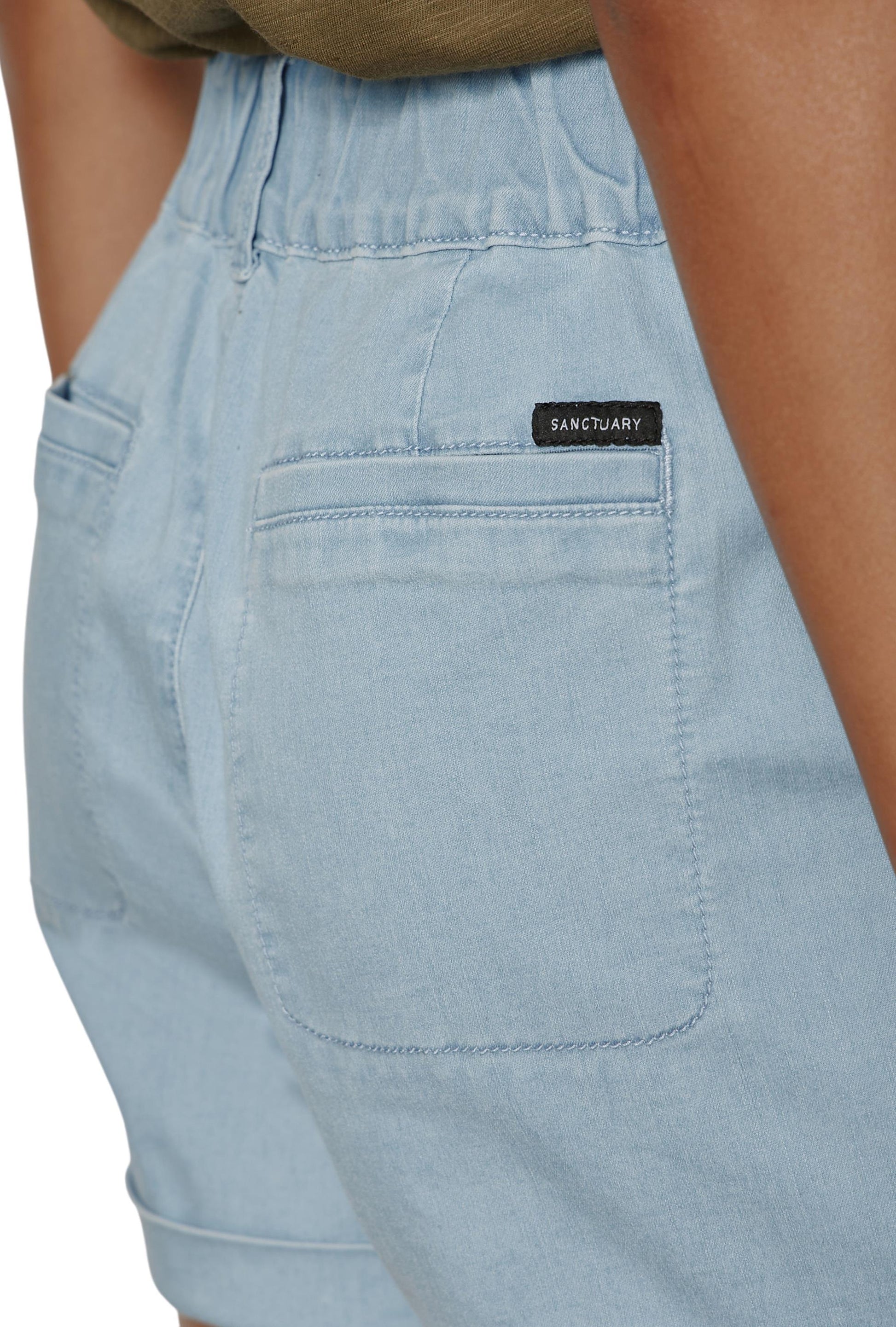 Close-up of a person wearing Sanctuary's Wander On Short in light blue denim, featuring an elastic waistband, back patch pockets, and a black Sanctuary label above the right pocket for added style and comfort.