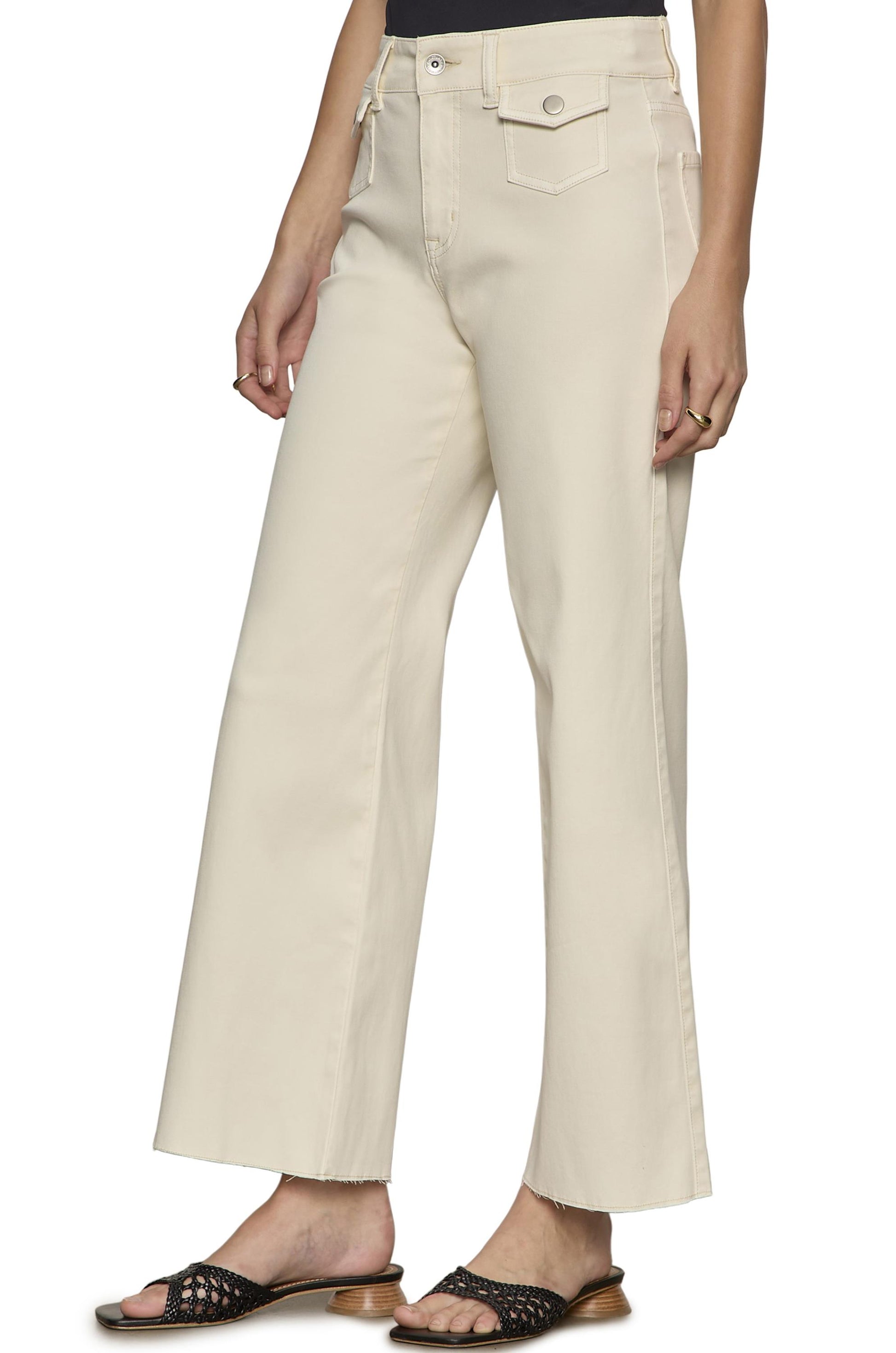 A person wearing Sanctuary Skipper Crop high-waisted, relaxed fit, light beige jeans with a front pocket and black woven sandals, shown from the waist down.