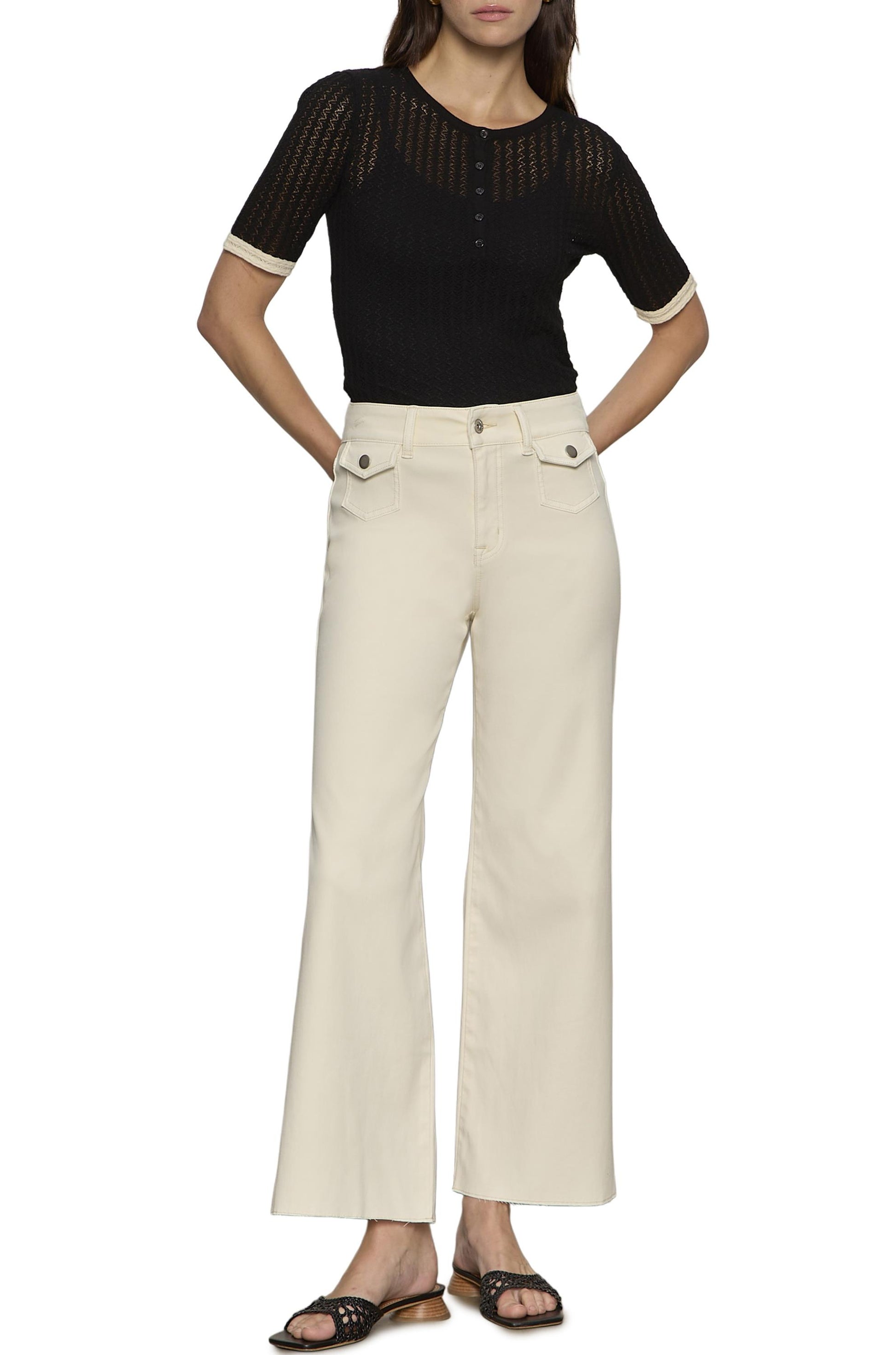 A woman stands with hands in her pockets, wearing Sanctuary's Skipper Crop high-waisted, wide-leg cream pants with front pockets, a black short-sleeve knit top, and black slide sandals.